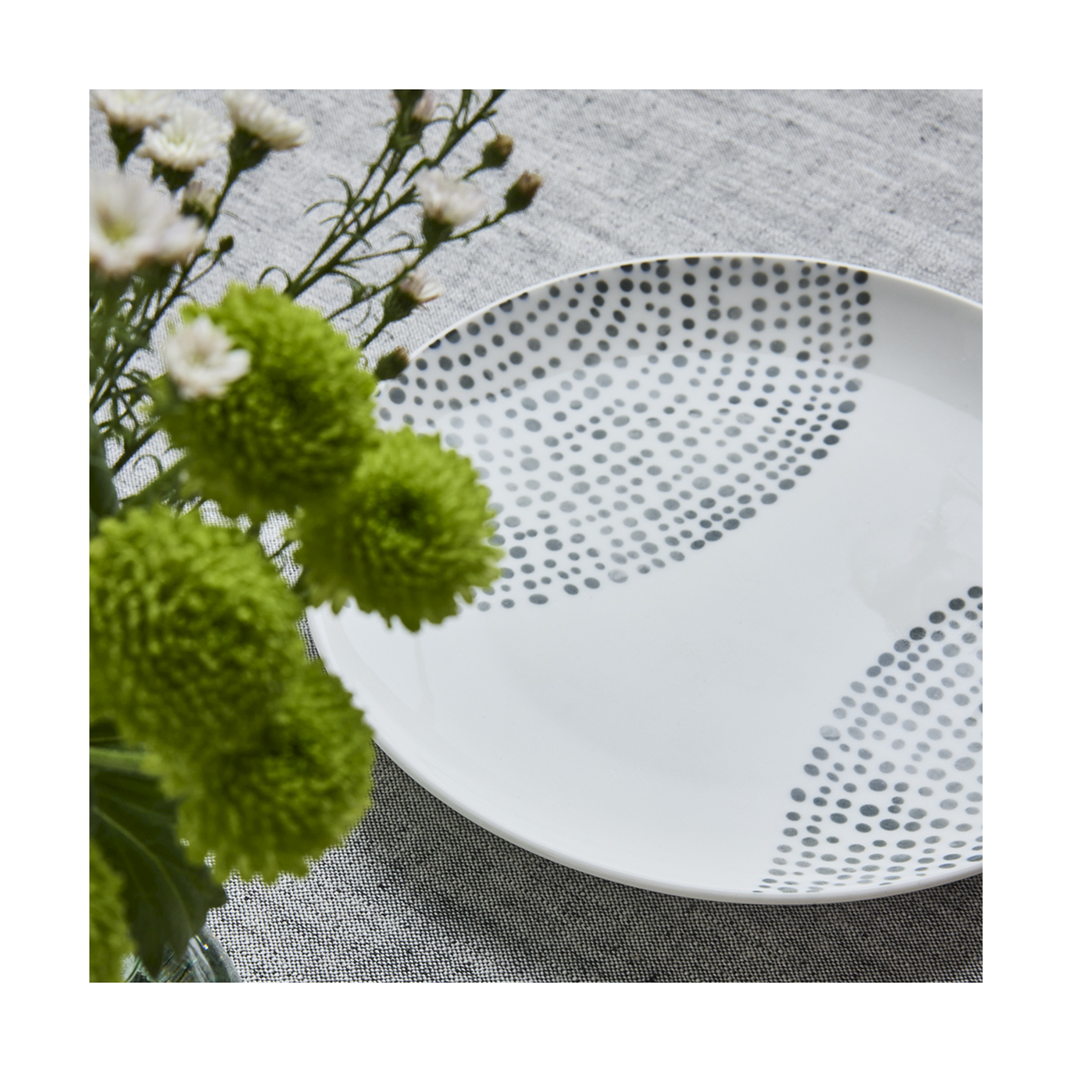 Un piatto bianco con pois grigi su un panno grigio testurizzato. Fiori verdi e bianchi a sinistra.