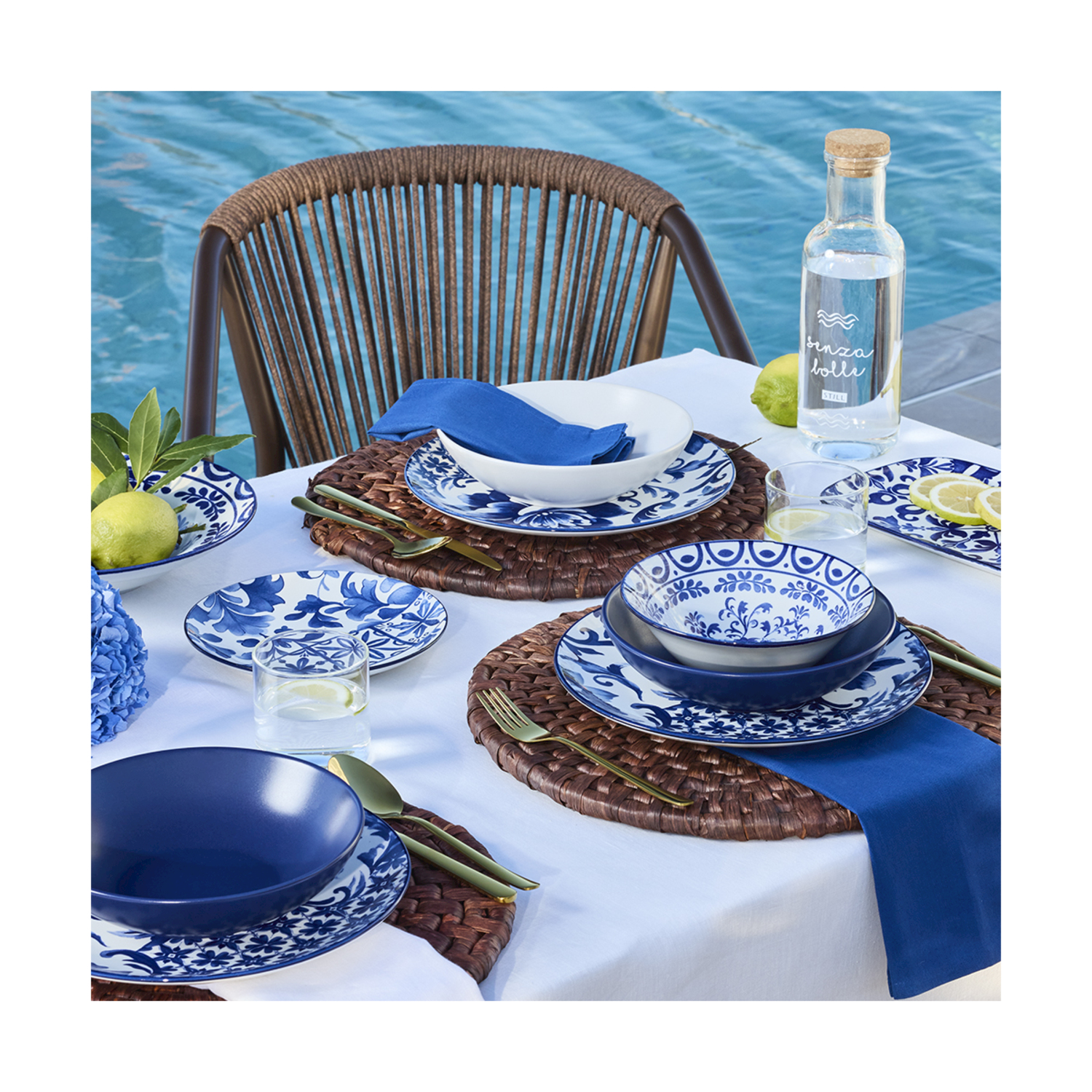 Una tavola apparecchiata con piatti blu e bianchi, posate dorate e limoni vicino a una piscina.