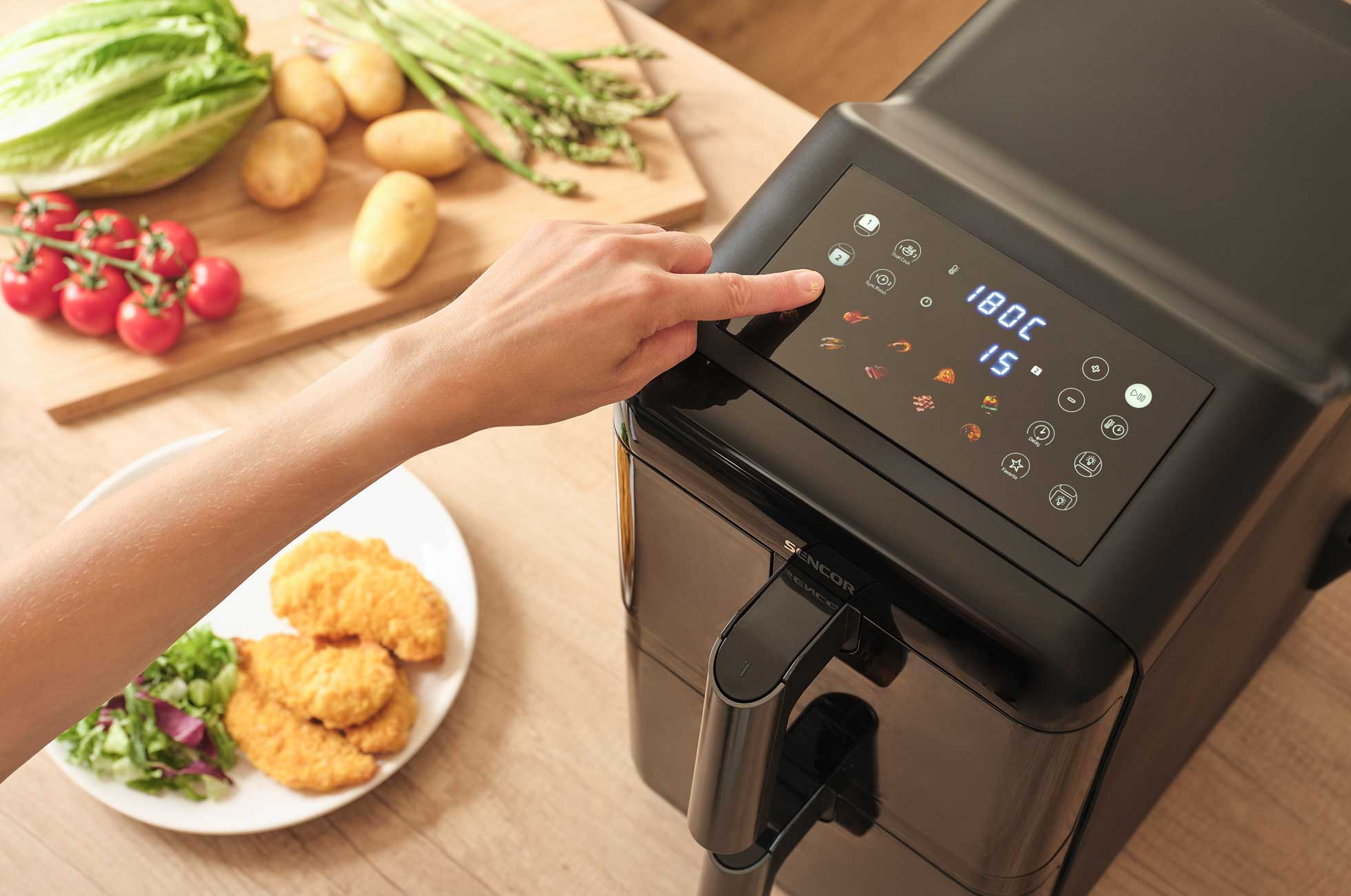 Egy kéz fekete, digitális kijelzős forrólevegős fritőz7t kezel. Mellette étel van.