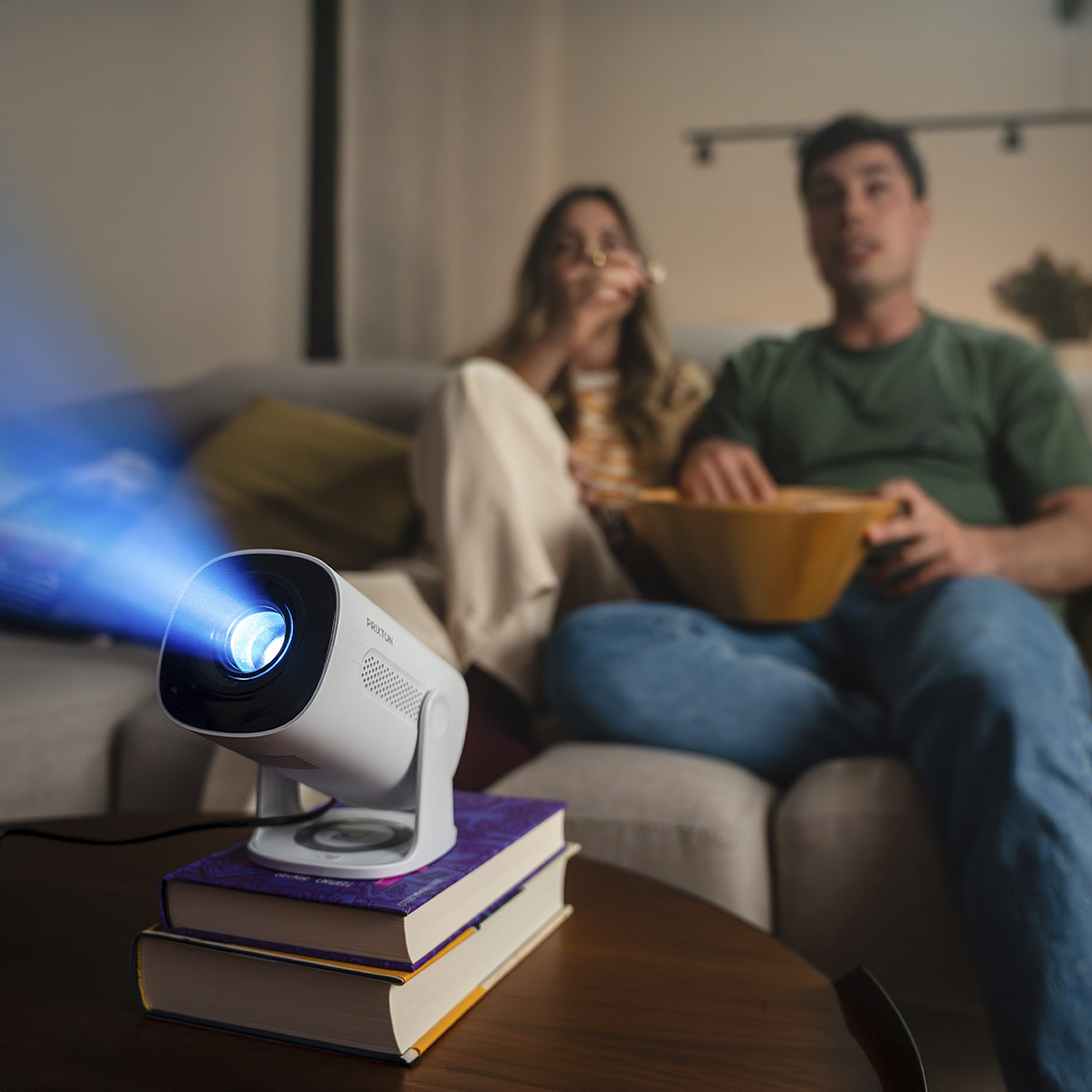 Un proyector blanco emite un haz de luz azul sobre libros, con una pareja sentada en un sofá.