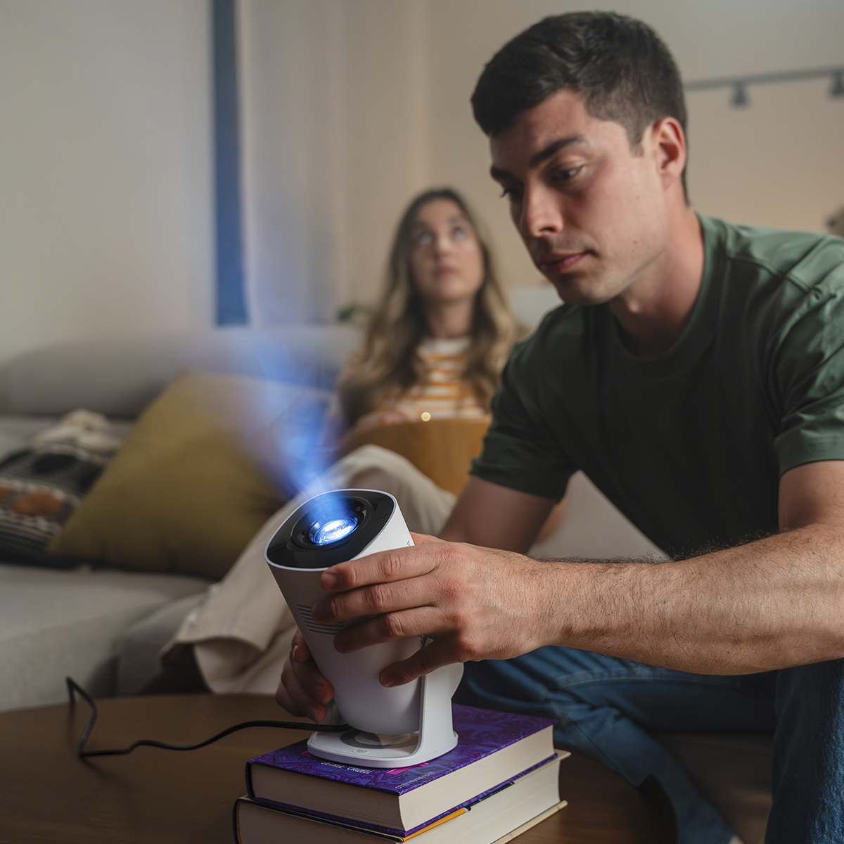 Hombre sostiene proyector emitiendo haz de luz azul, mujer sentada detrás.