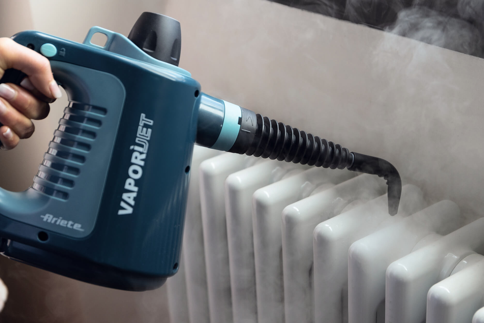 Una mano sostiene un limpiador a vapor azul apuntado a un radiador blanco, con vapor saliendo.
