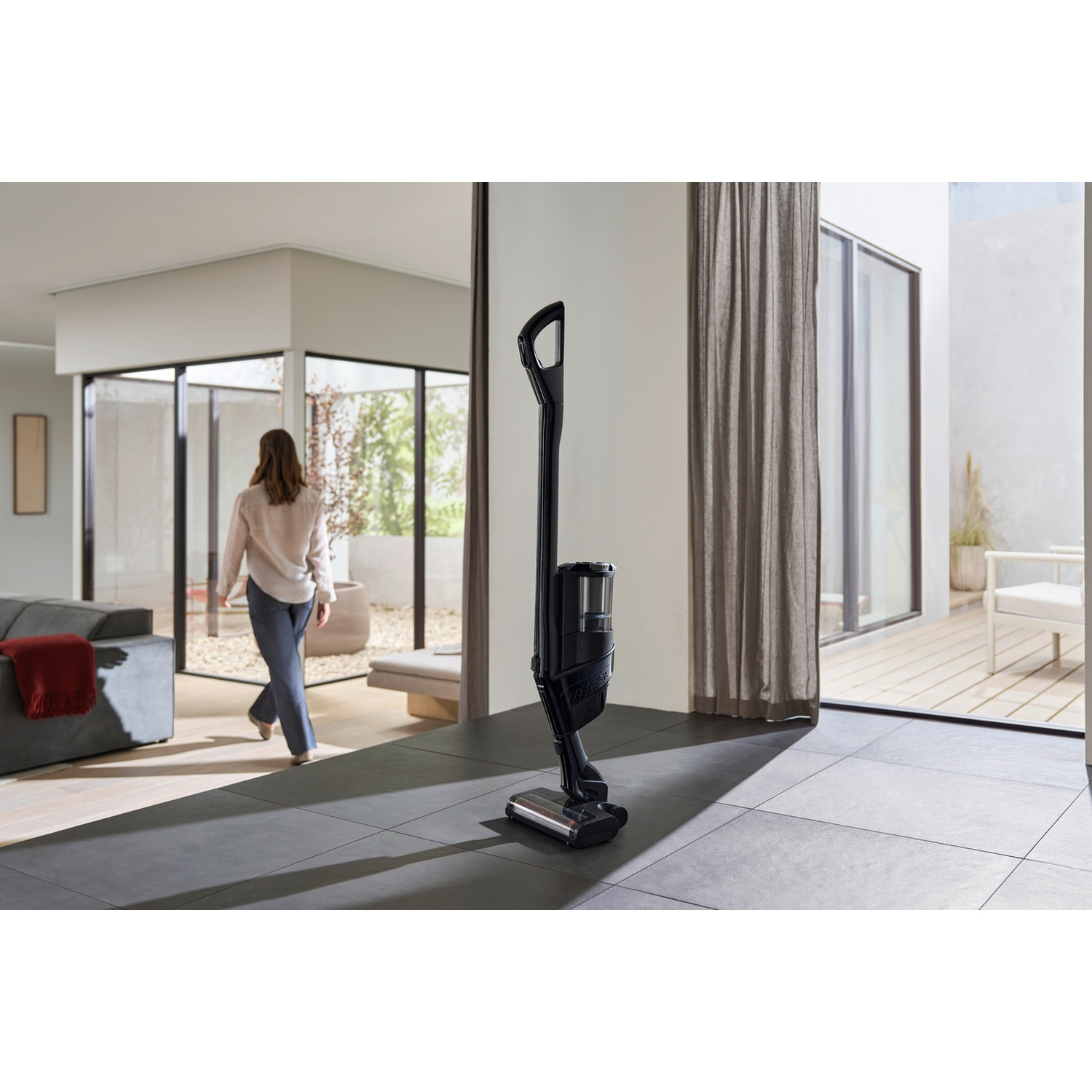 A black cordless vacuum stands on a gray tiled floor. A woman walks past.