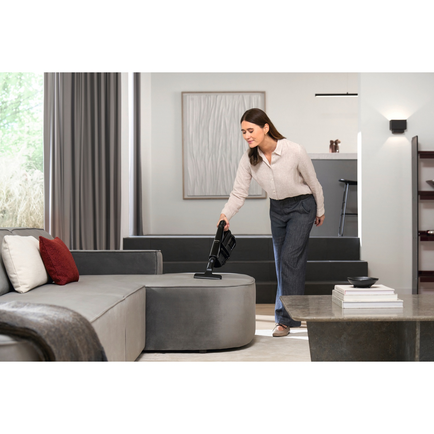 A woman vacuums a gray ottoman in a modern living room with a sofa.