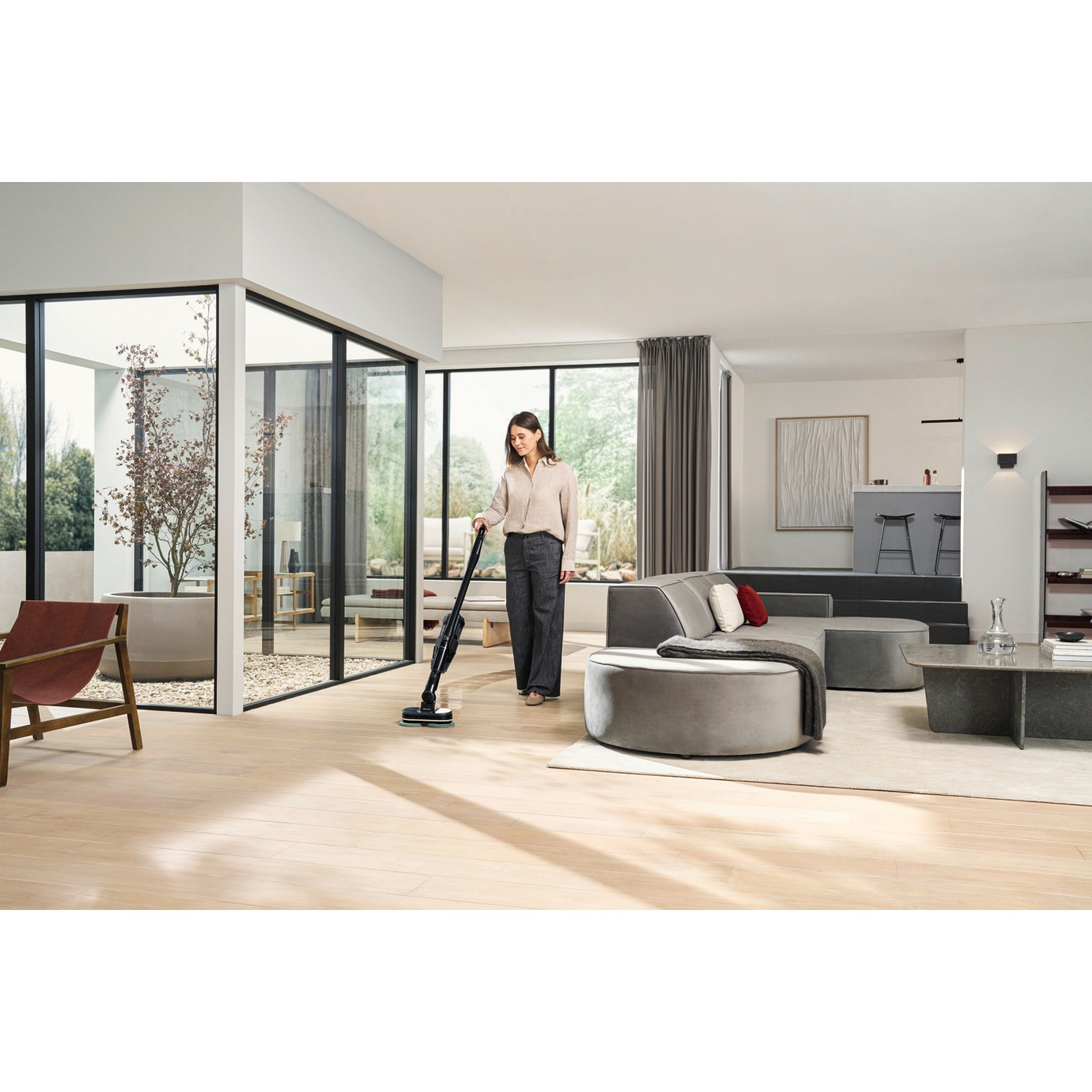 A woman vacuums a light wood floor in a modern living room with large windows.