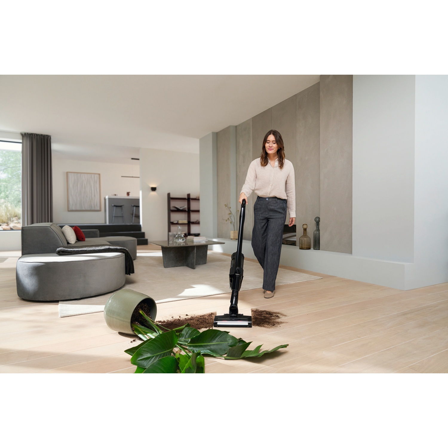 Femme passe l'aspirateur sur une plante renversée et de la terre dans un salon moderne.