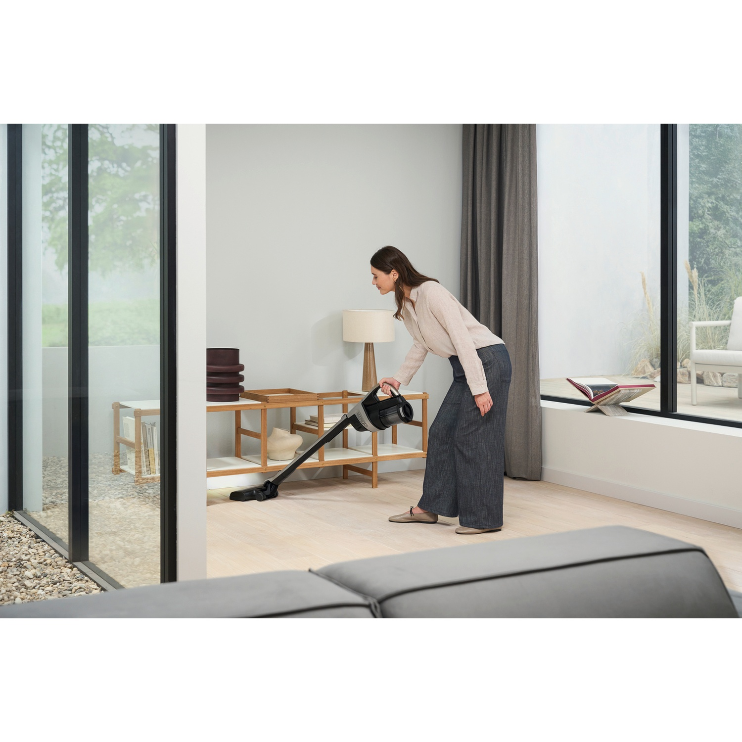 Une femme passe l'aspirateur sur un parquet clair avec un aspirateur sans fil noir.