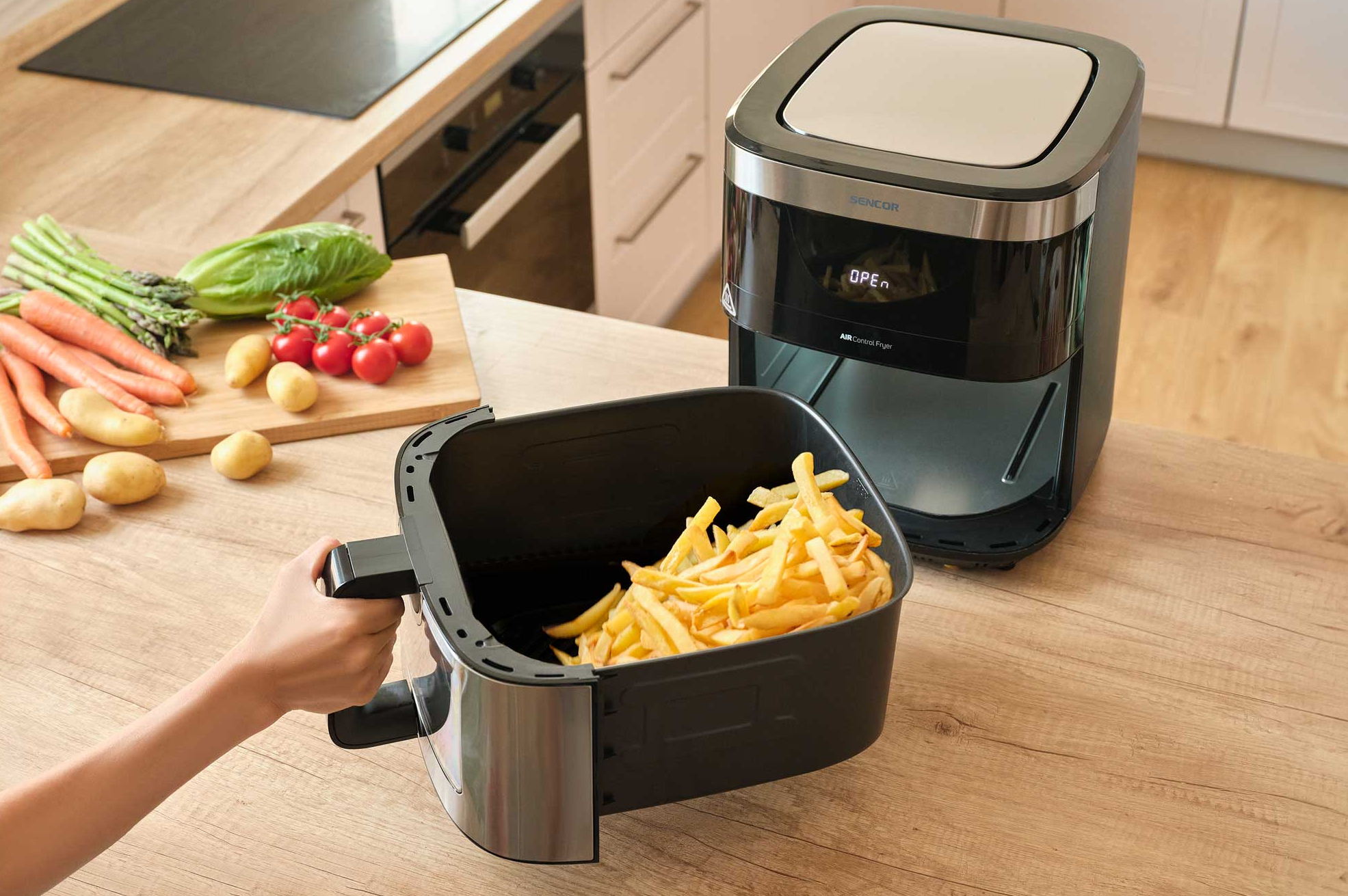 Egy kéz sültkrumplival teli air fryer kosarat tart a fritő mellet.