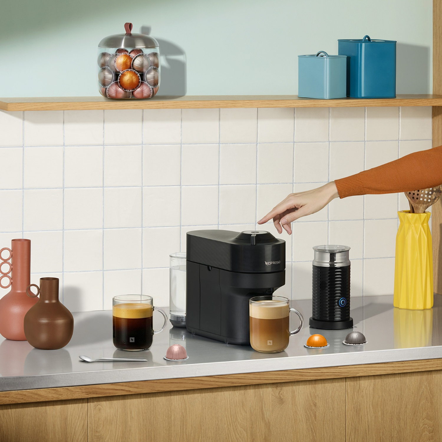 A black Nespresso machine with coffee, frother, two vases, and capsules on a counter.