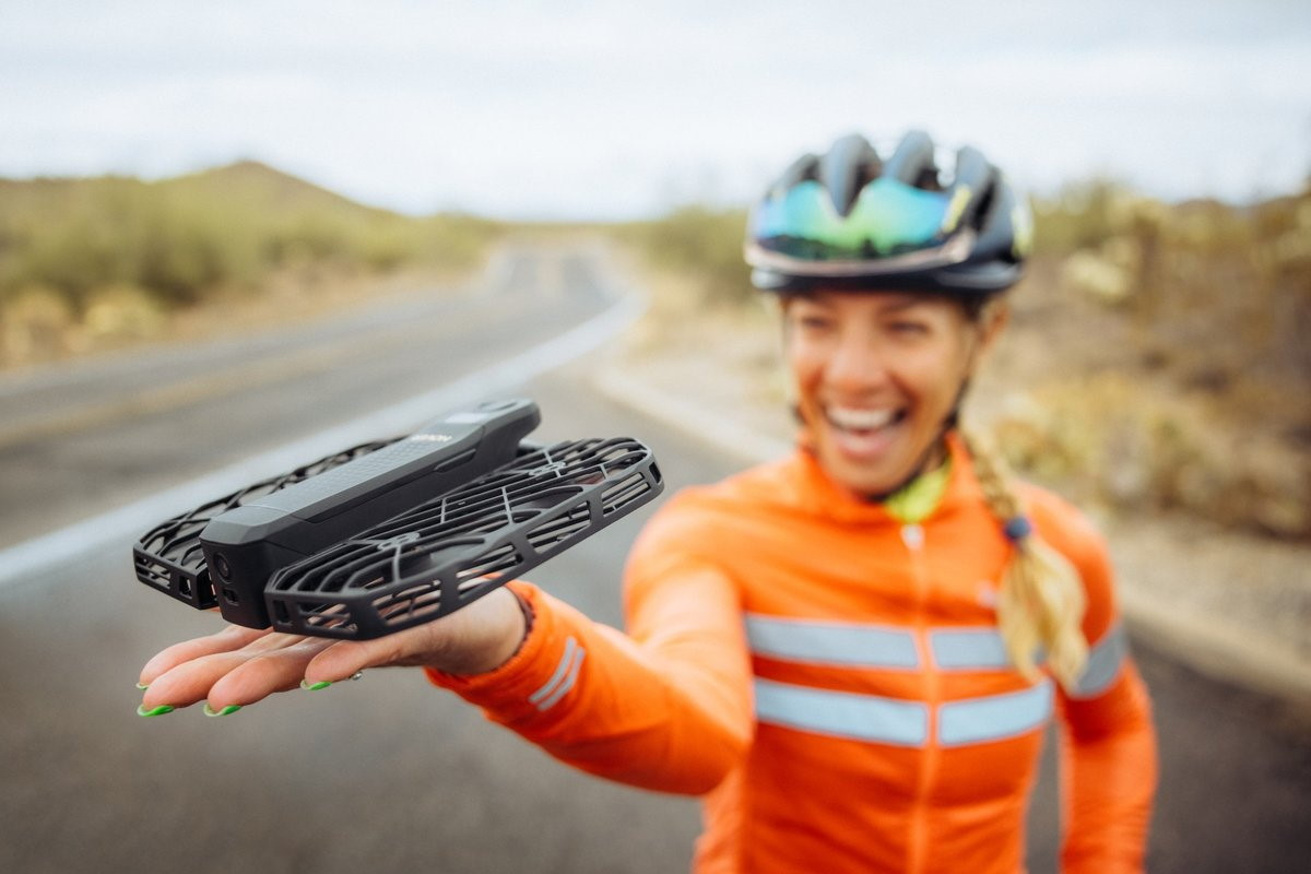 Una ciclista tiene un drone in mano. Indossa una tuta da ciclismo arancione.