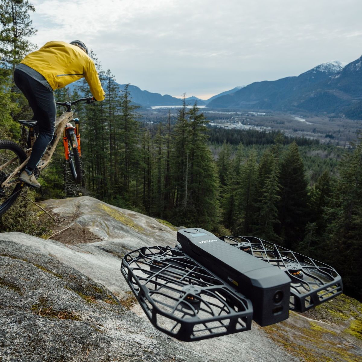 Un mountain biker salta su rocce con foresta e montagne sullo sfondo. Un drone è in primo piano.