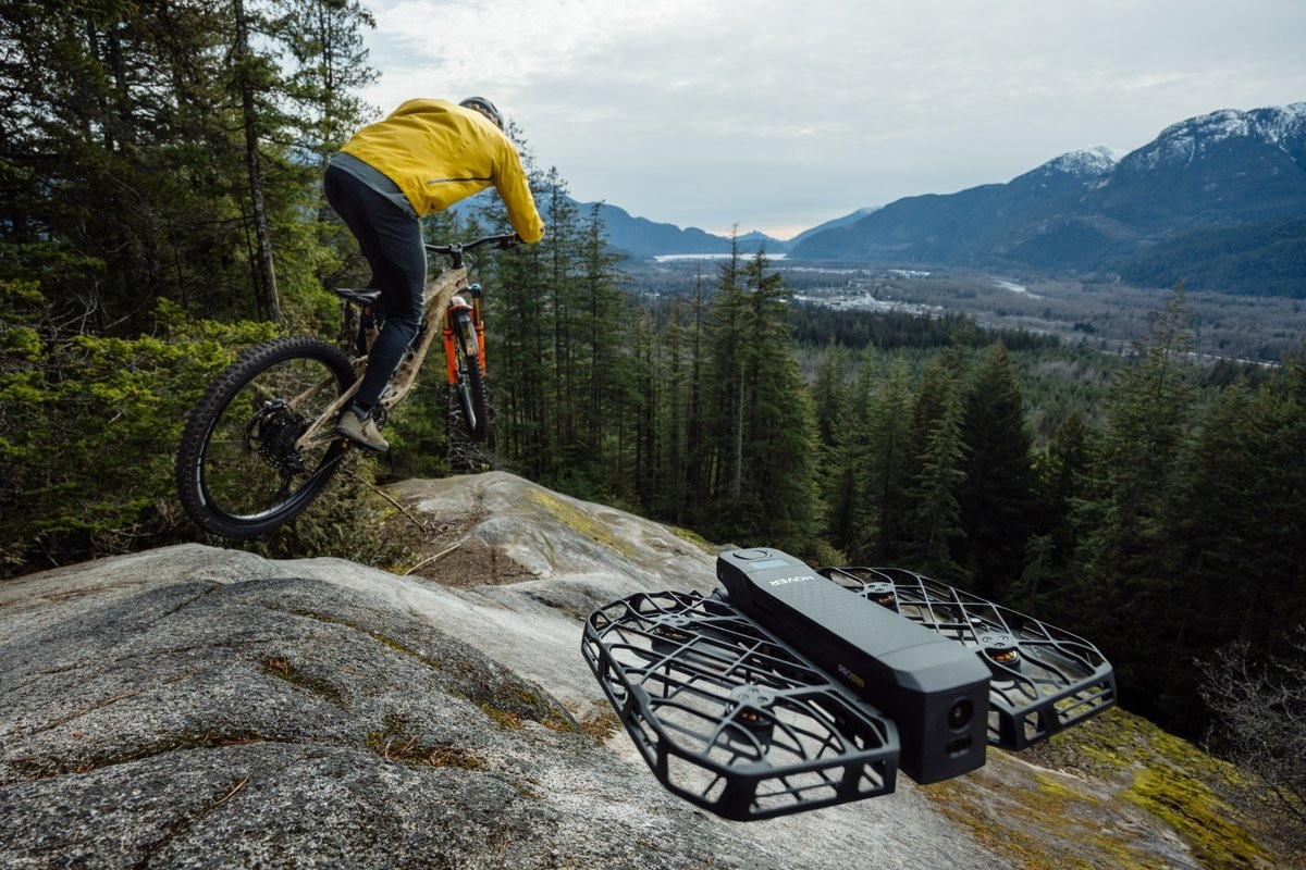 Un ciclista in giacca gialla salta su rocce con un drone in primo piano.