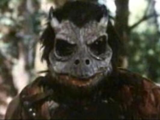 Ein Wesen mit einer tiermaskenartigen Fratze und dunklem Fell steht im Wald.