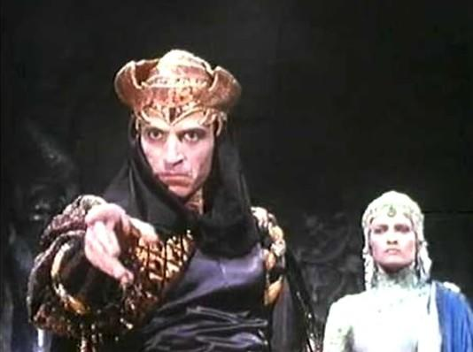 Ein Mann mit goldenem Helm und schwarzem Umhang zeigt nach vorne. Eine Frau mit Kopfschmuck steht daneben.