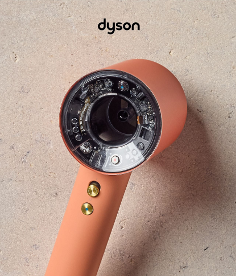 Koralowy suszarka do włosów Dyson z widoczną elektroniką i złotymi przyciskami.