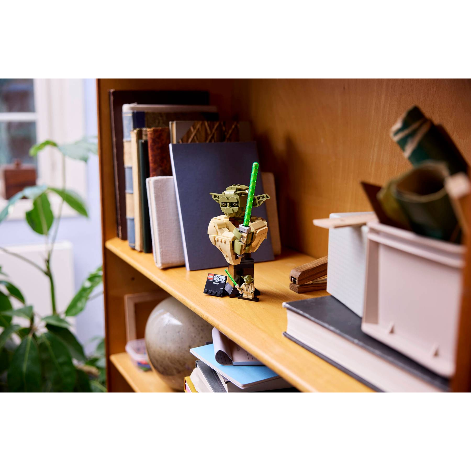 A Lego Yoda figure holding a lightsaber stands on a bookshelf next to books.