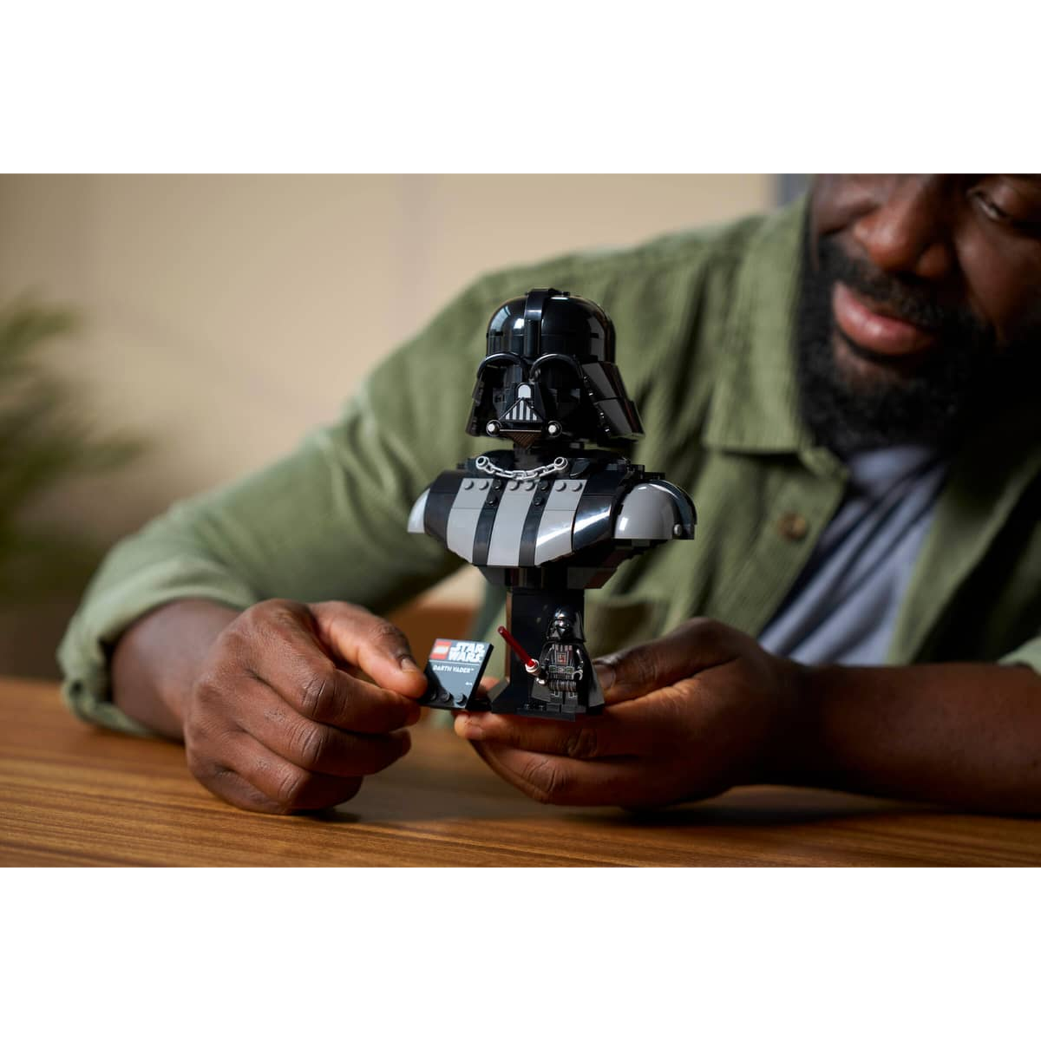 Man in green shirt builds LEGO Darth Vader bust, holding nameplate.