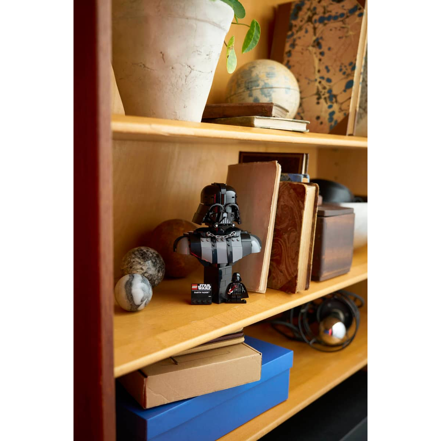A Darth Vader Lego bust on a shelf with books and spheres.