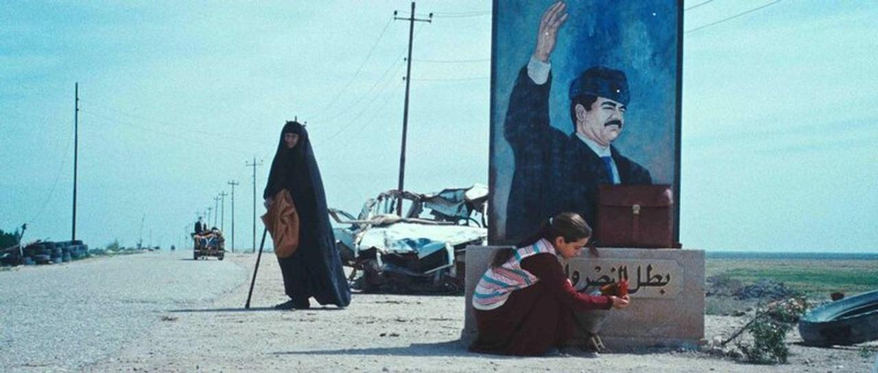 Ein Mädchen kniet vor einem Porträt von Saddam Hussein. Eine Frau in Schwarz geht vorbei.