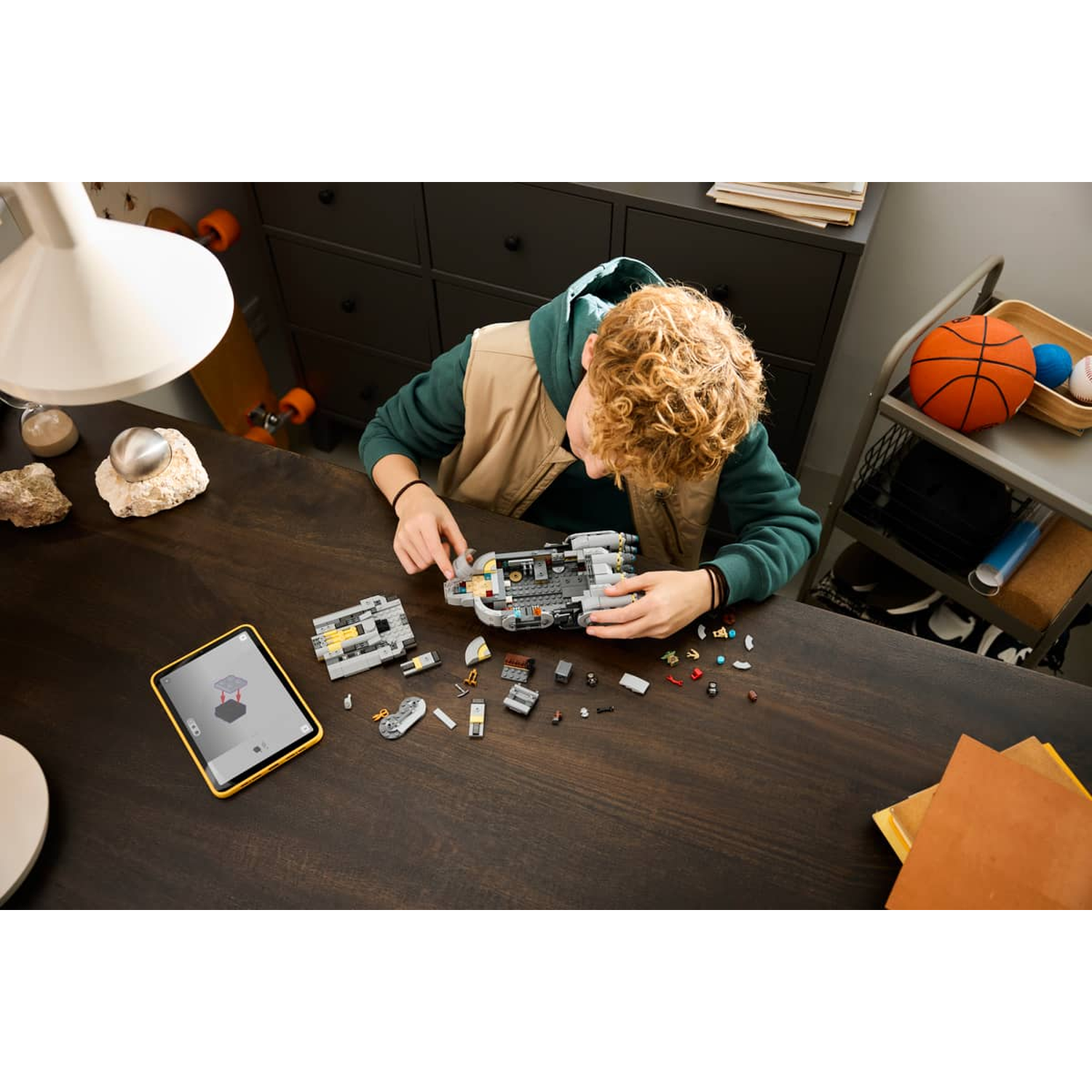A boy builds a spaceship from Lego bricks on a wooden table, with instructions on a tablet.