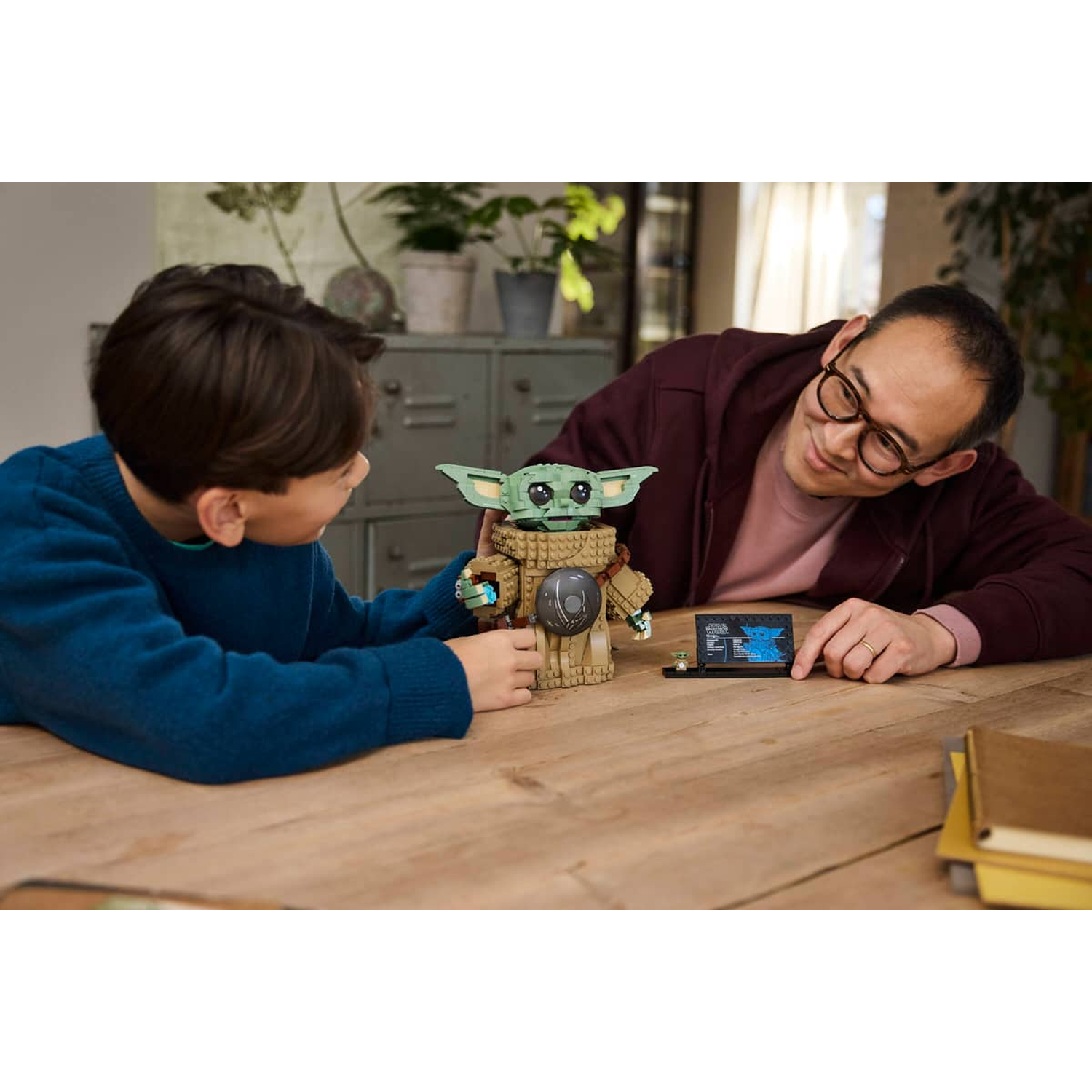 Een man en een jongen bouwen een Lego-model van Baby Yoda aan een tafel.