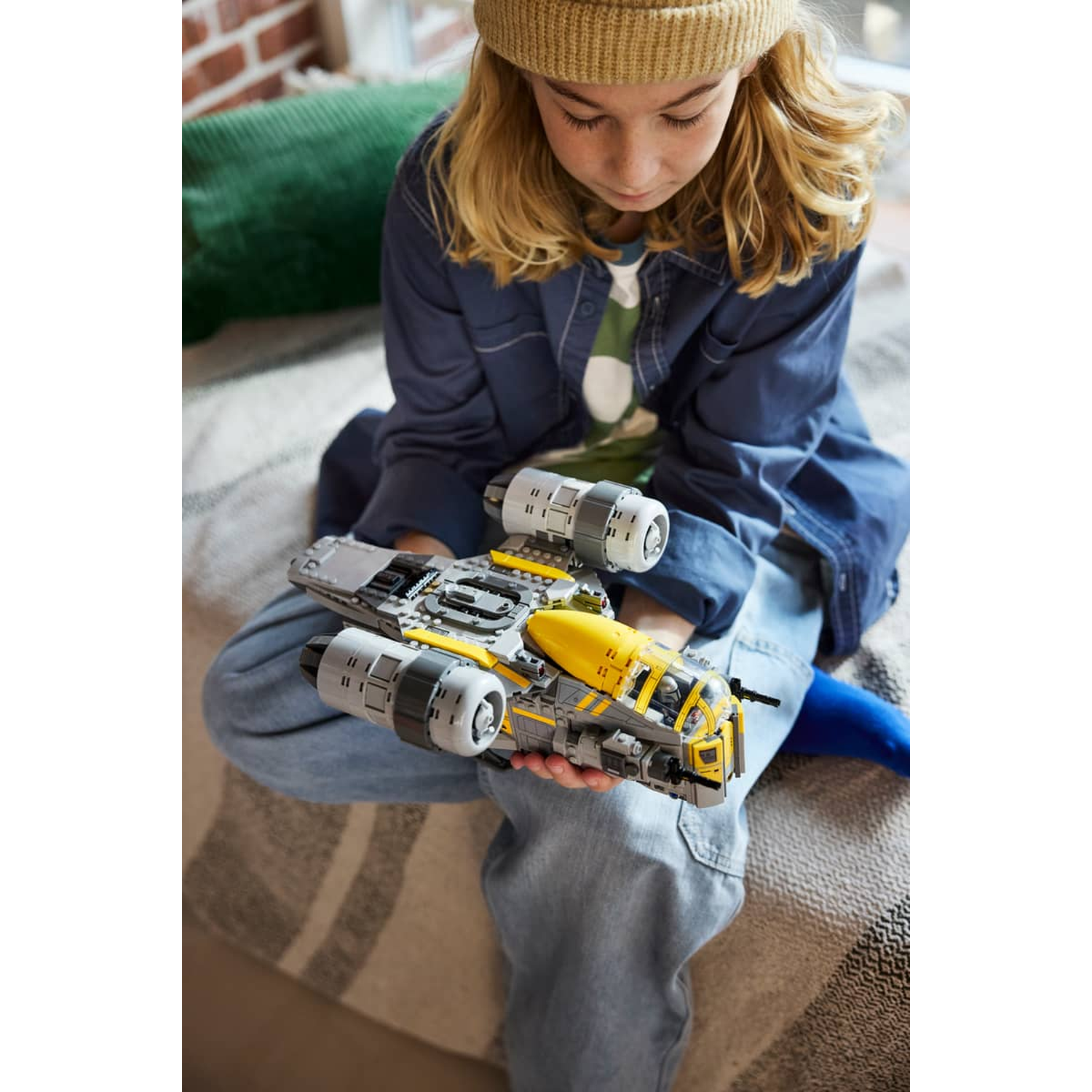 Child wearing a yellow beanie holds a gray and yellow toy spaceship.