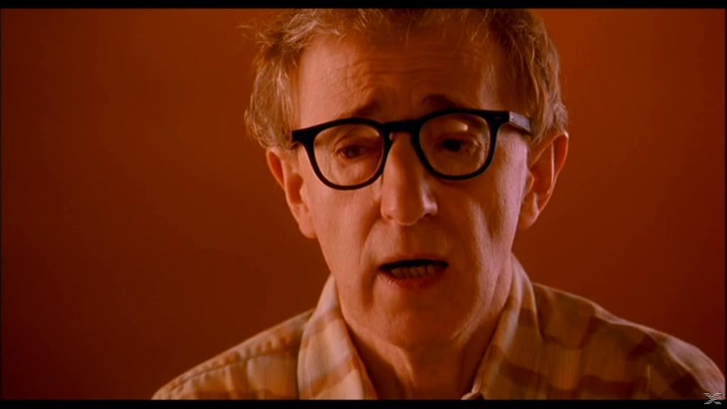 Ein Mann mit Brille und kariertem Hemd spricht vor einem orangefarbenen Hintergrund.