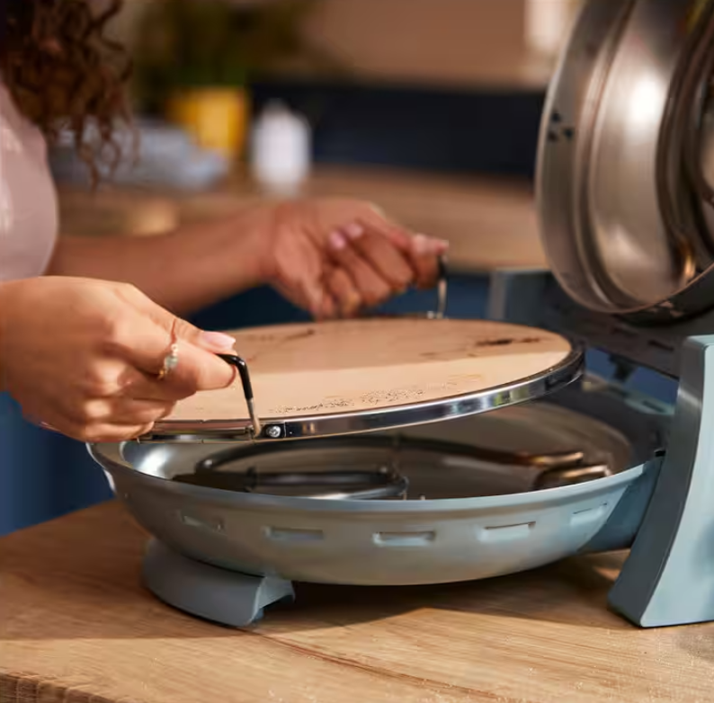 Mani che sollevano una pietra rotonda per pizza con manici neri da un forno blu chiaro.