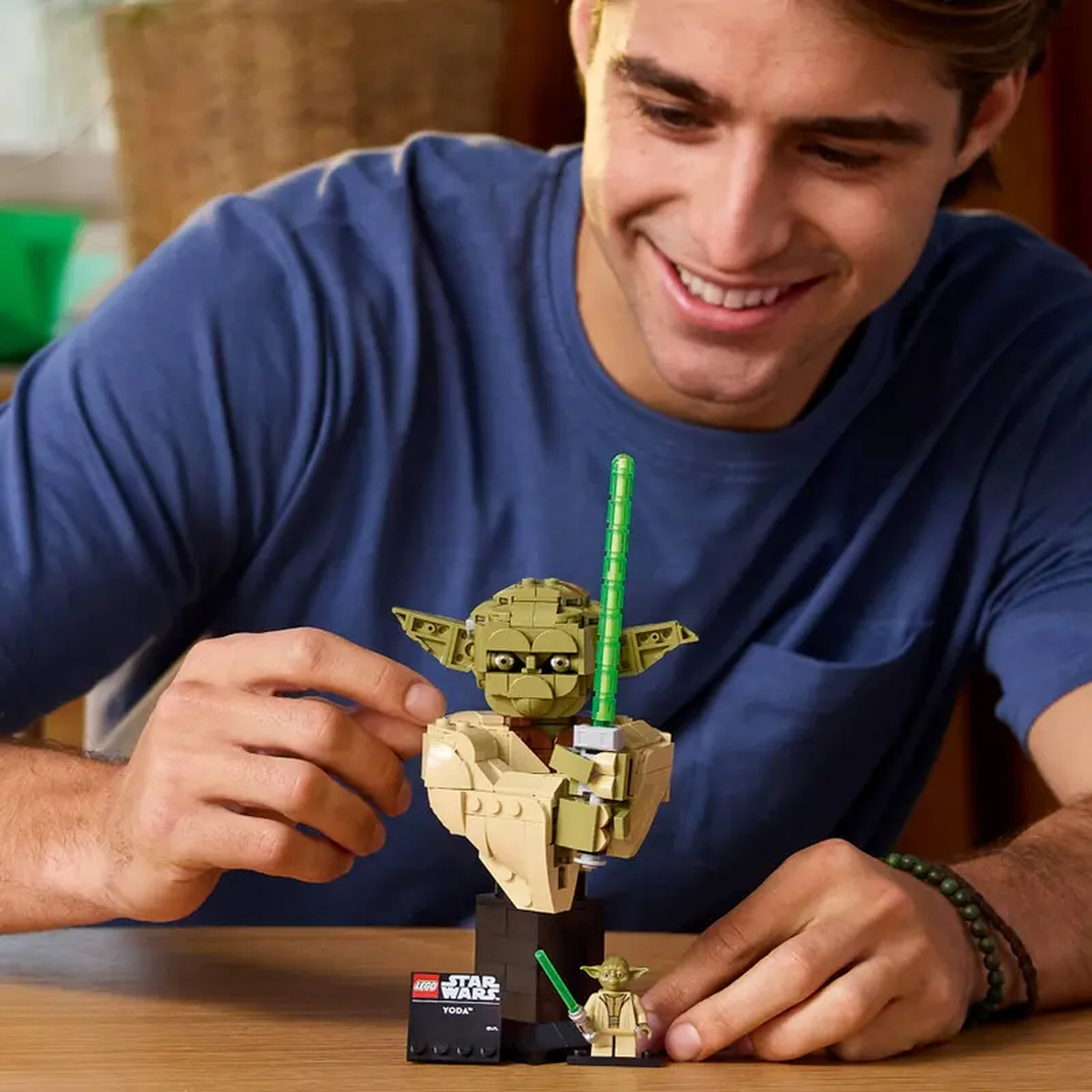 A smiling man builds a Lego Yoda bust and mini-figure with green lightsabers.