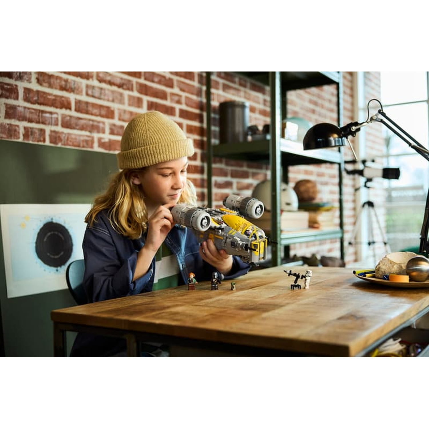 A girl wearing a yellow beanie builds a Lego spaceship on a wooden table.