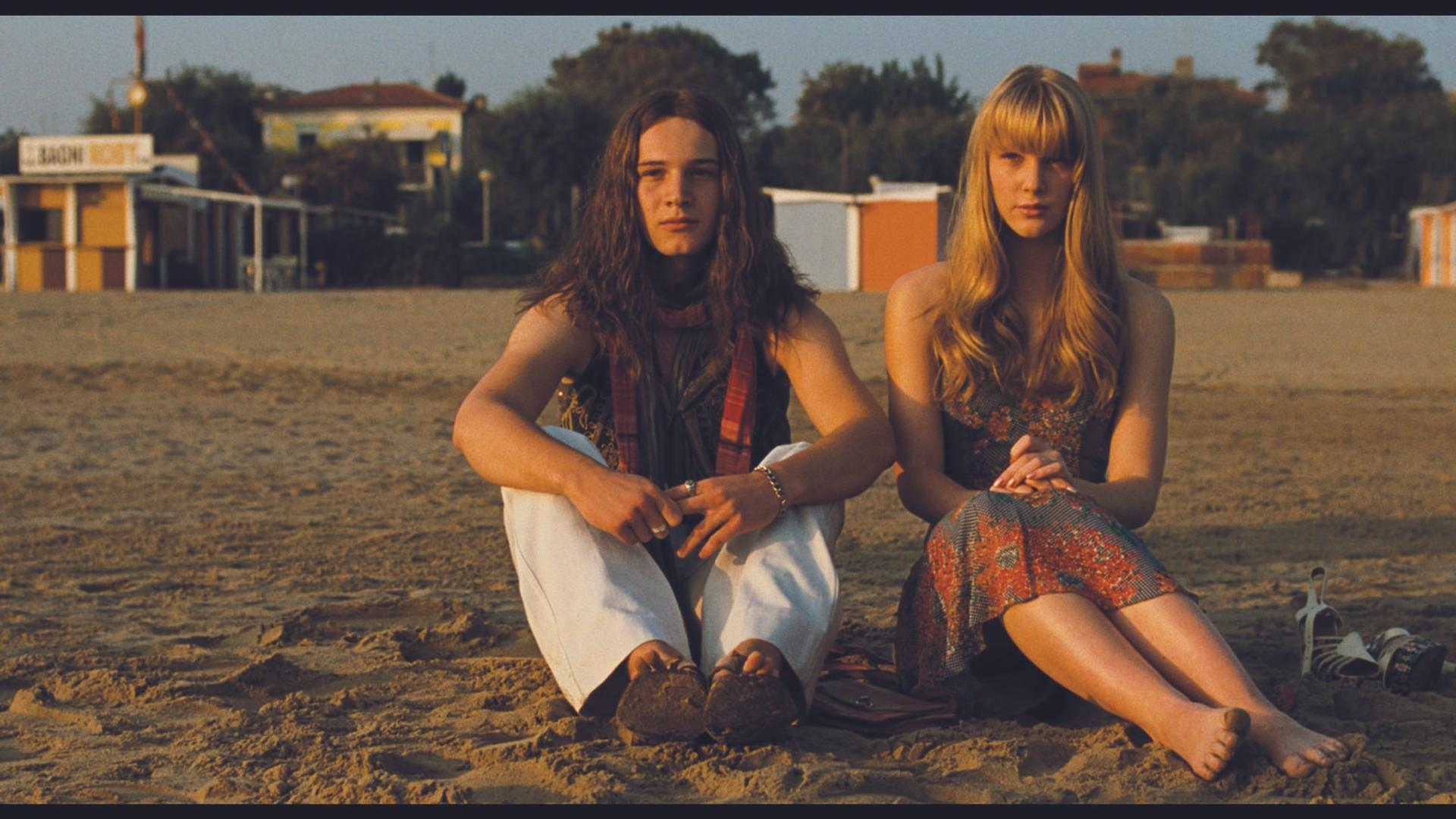Ein Mann mit langen Haaren und eine Frau sitzen am Strand. Im Hintergrund sind Gebäude.