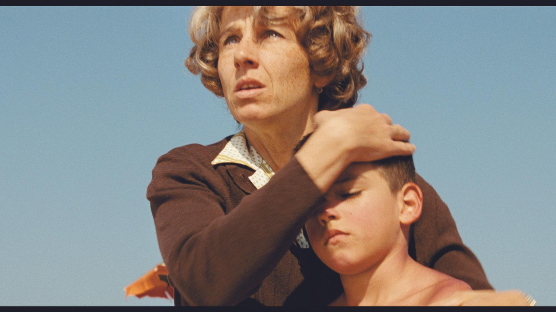Eine Frau legt ihre Hand auf den Kopf eines Jungen vor blauem Himmel.