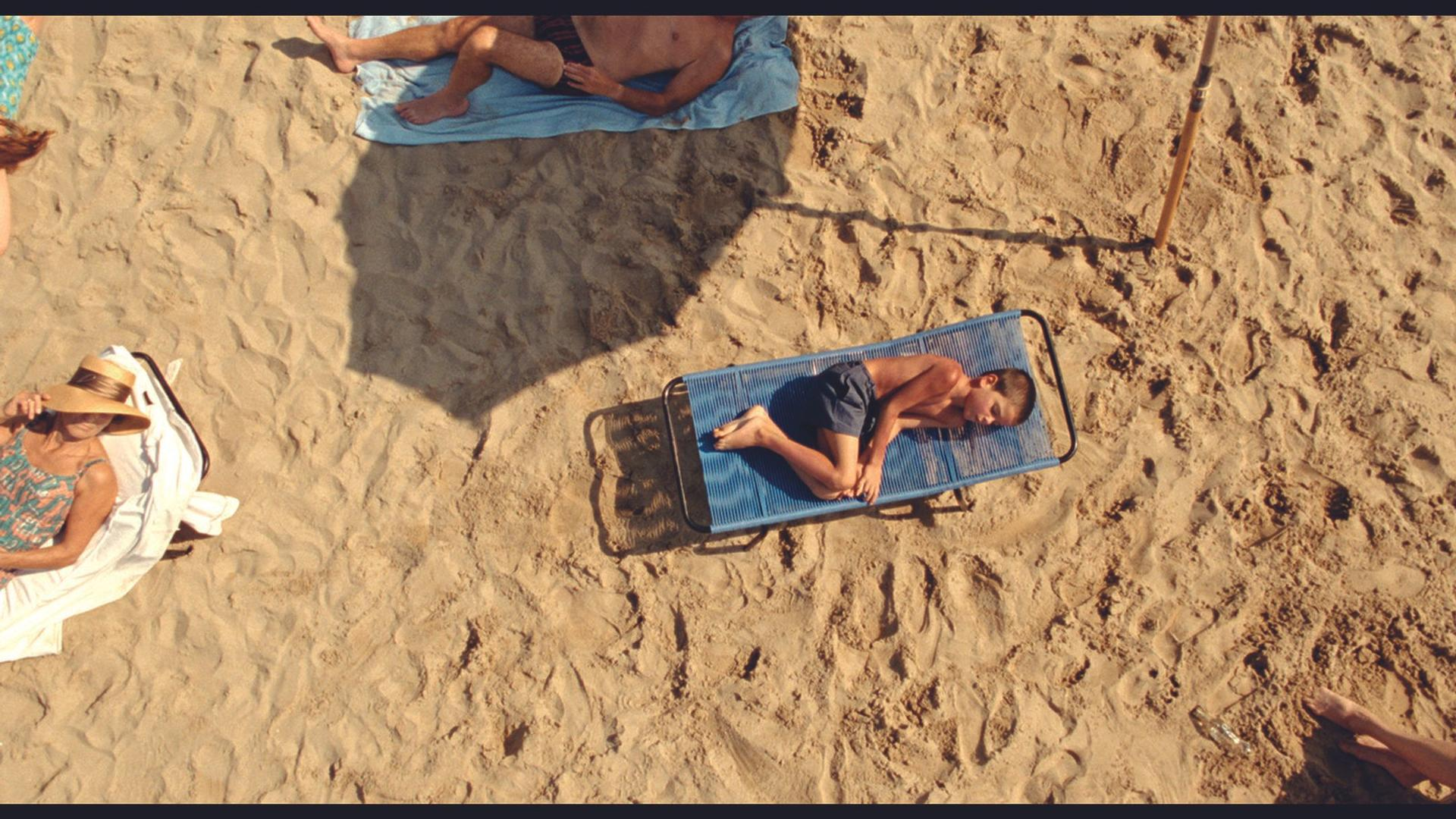 Ein Junge schläft auf einer blauen Liege am Strand, während andere sich sonnen.
