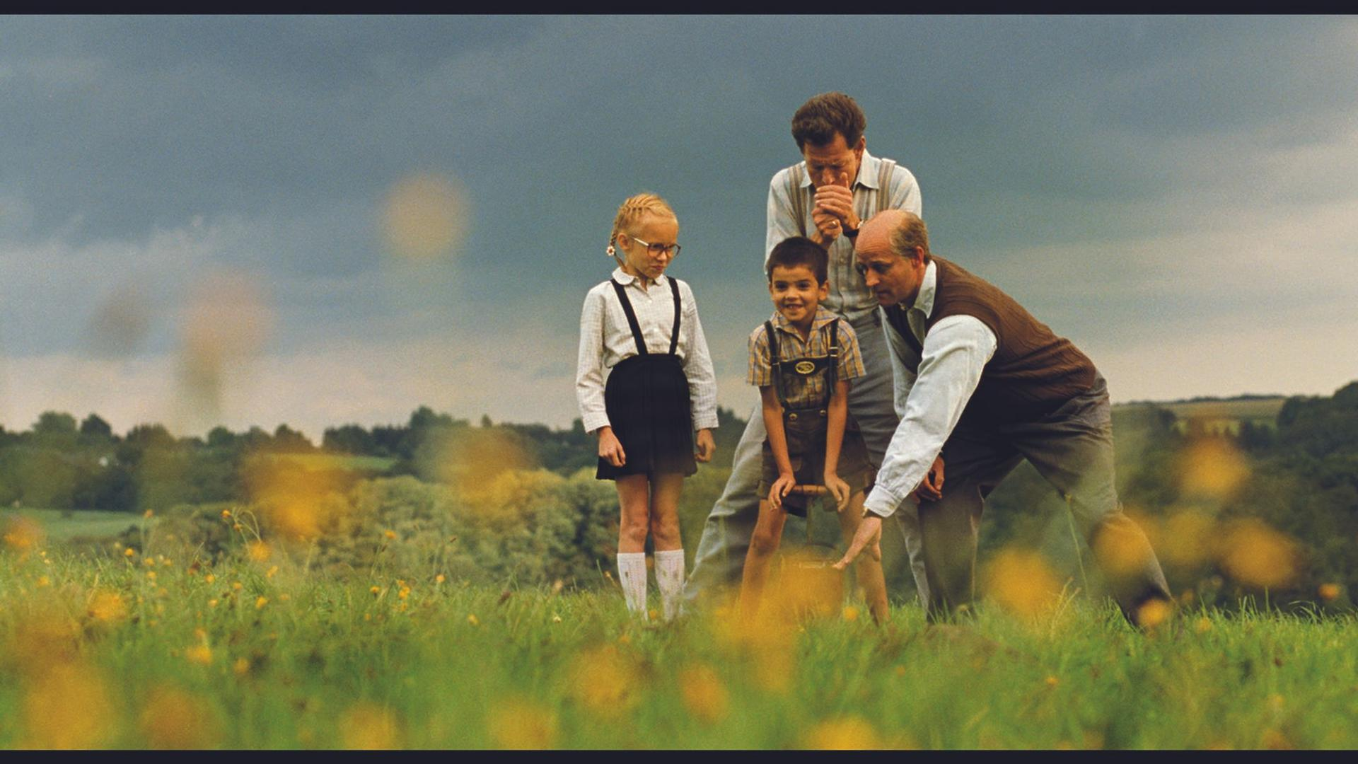 Zwei Männer und zwei Kinder stehen auf einem Feld mit gelben Blumen unter einem dramatischen Himmel.