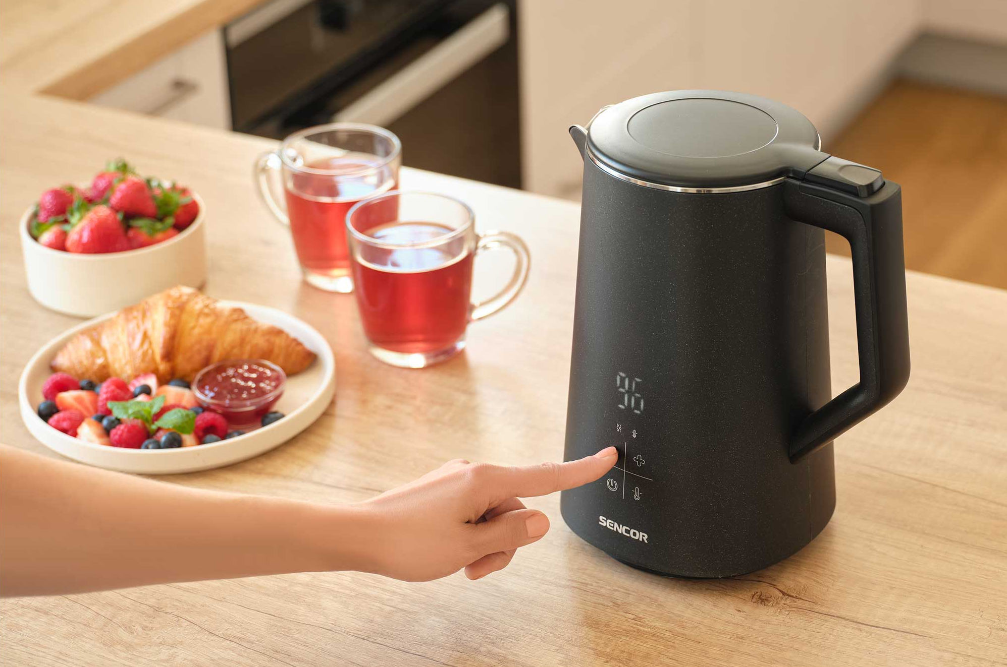 Egy kéz egy fekete, digitális kijelzős vízforralót nyom meg. Közelben két csésze tea és egy gyümölcsös, péksüteményes tányér látható.
