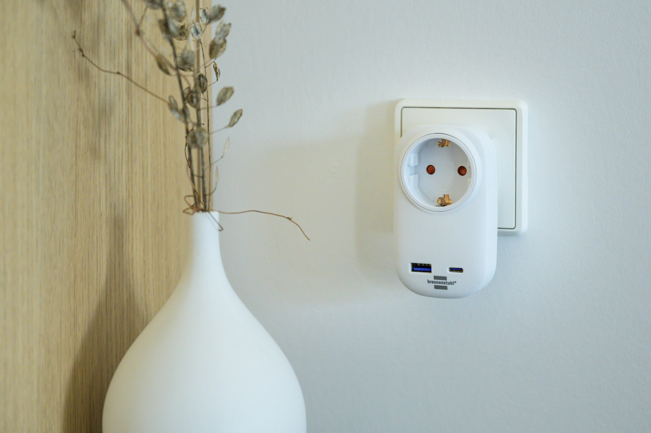 Ein weißer Steckeradapter mit USB-Anschlüssen, neben einer Vase mit Trockenblumen.