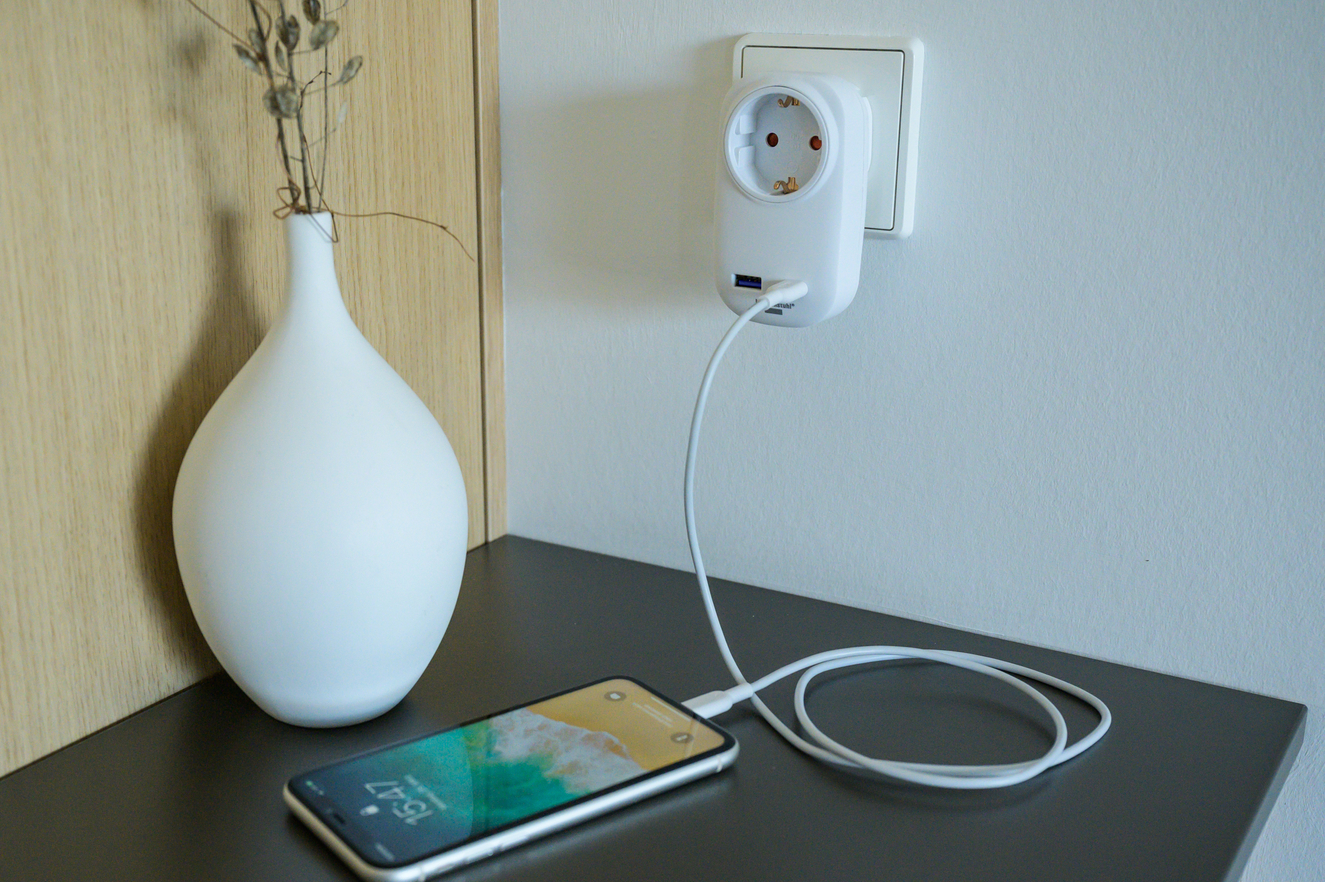 Ein weißer Steckeradapter mit USB-Anschluss lädt ein Smartphone auf dem Nachttisch.