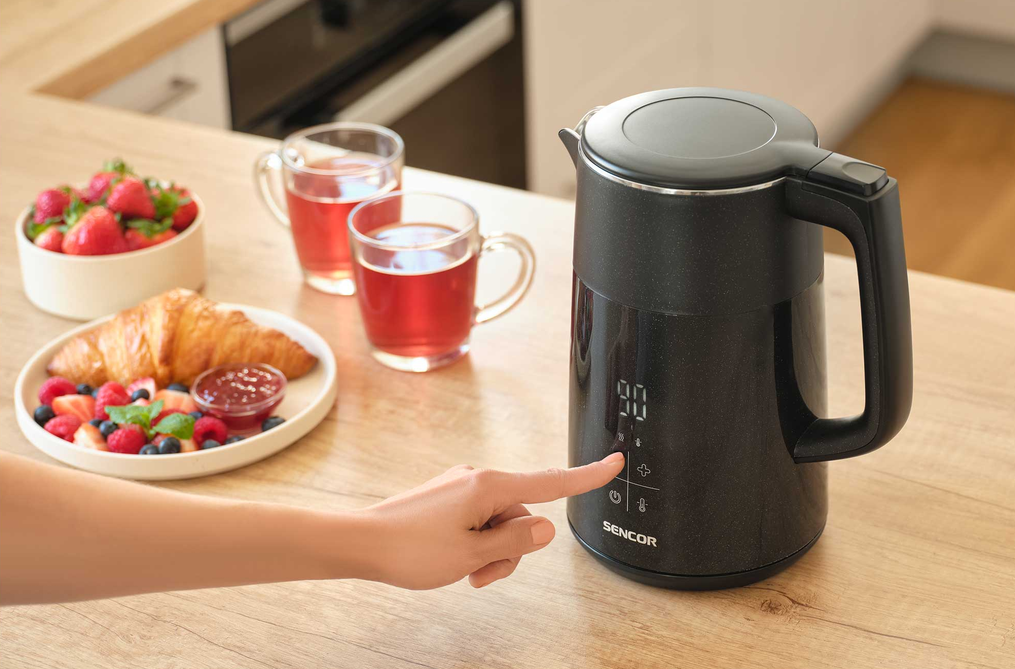Egy kéz fekete vízforralóra nyom, mellette reggeli bogyókkal és croissanttal.