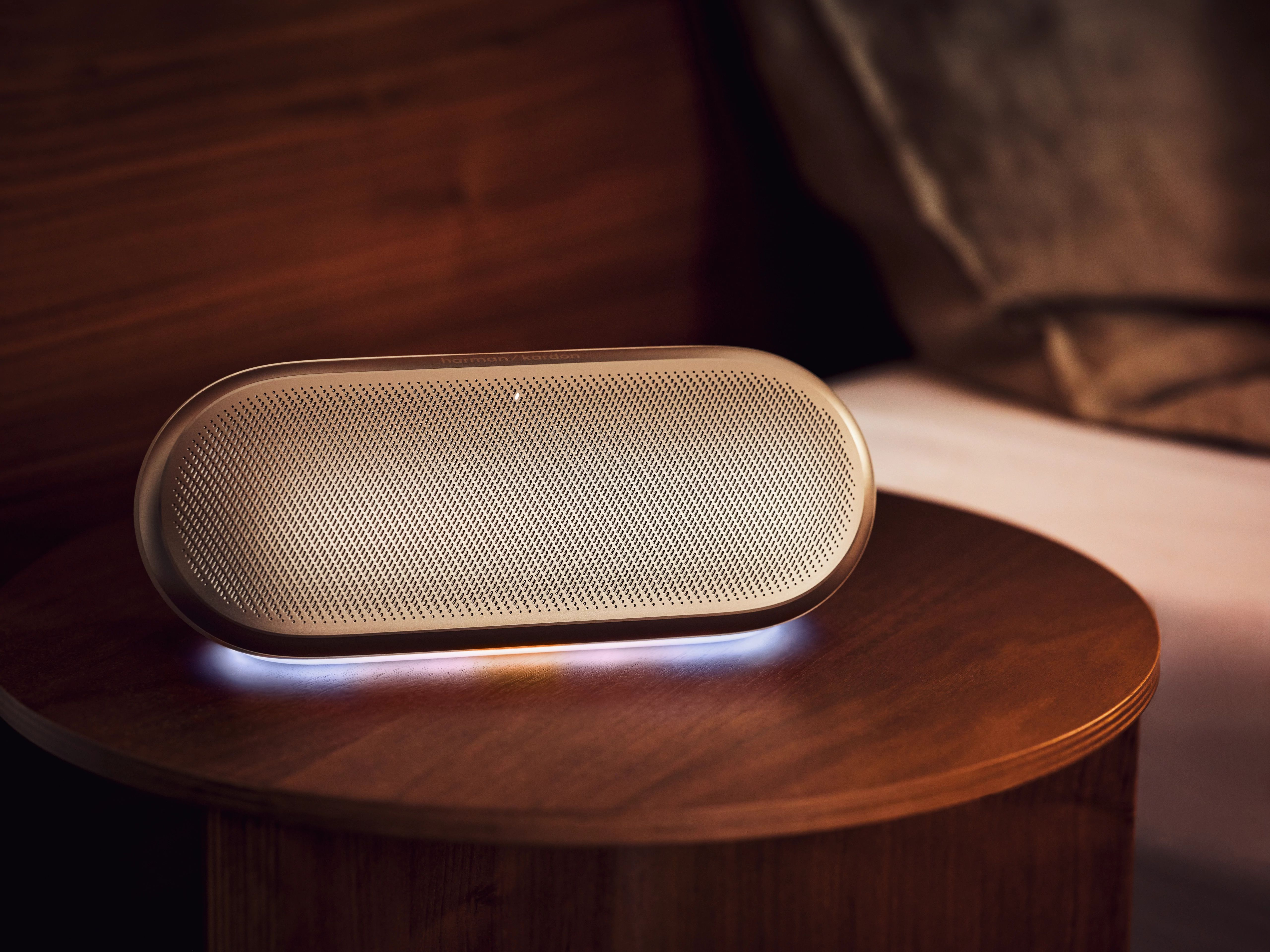 Ahşap bir masanın üzerinde aydınlatılmış altın rengi Harman Kardon hoparlör.