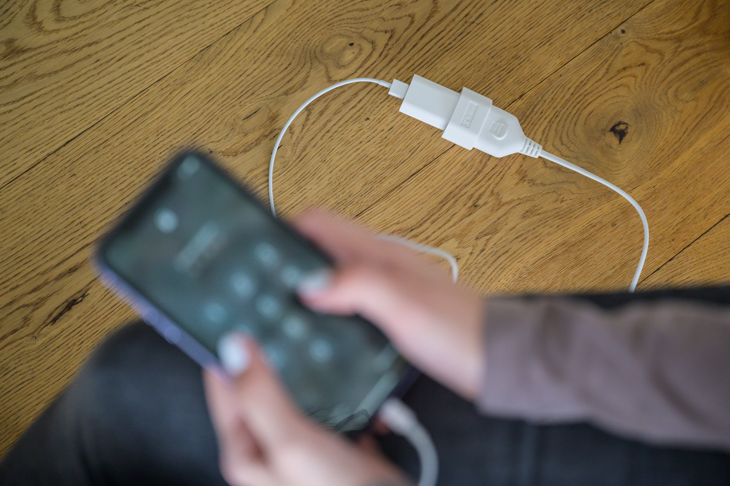 Eine Hand hält ein Smartphone, das an ein Ladegerät angeschlossen ist, auf Holzboden.