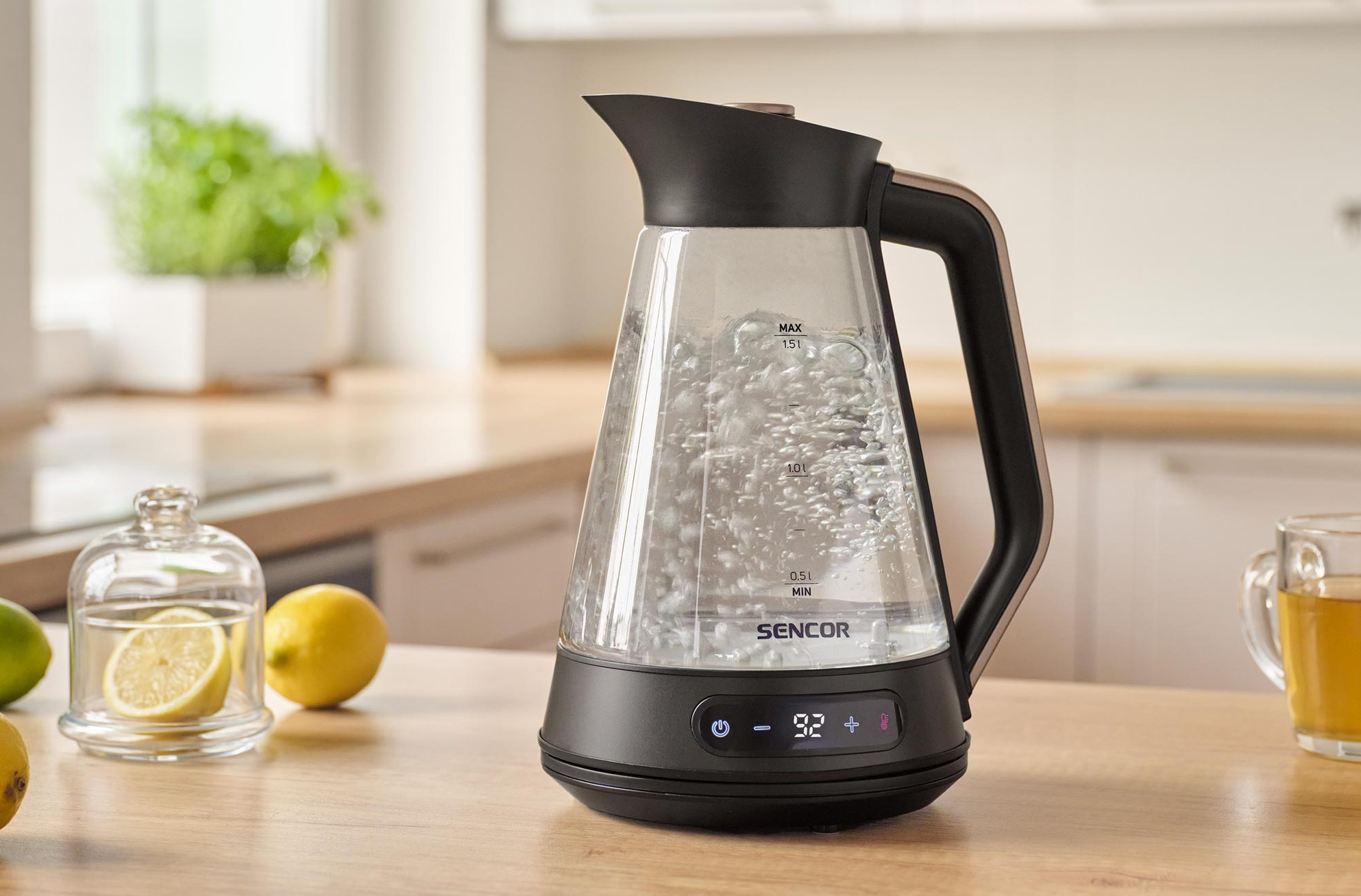 Egy üveg vízforralóban forr a víz buborékokkal. Citromok és egy csésze tea mellette.