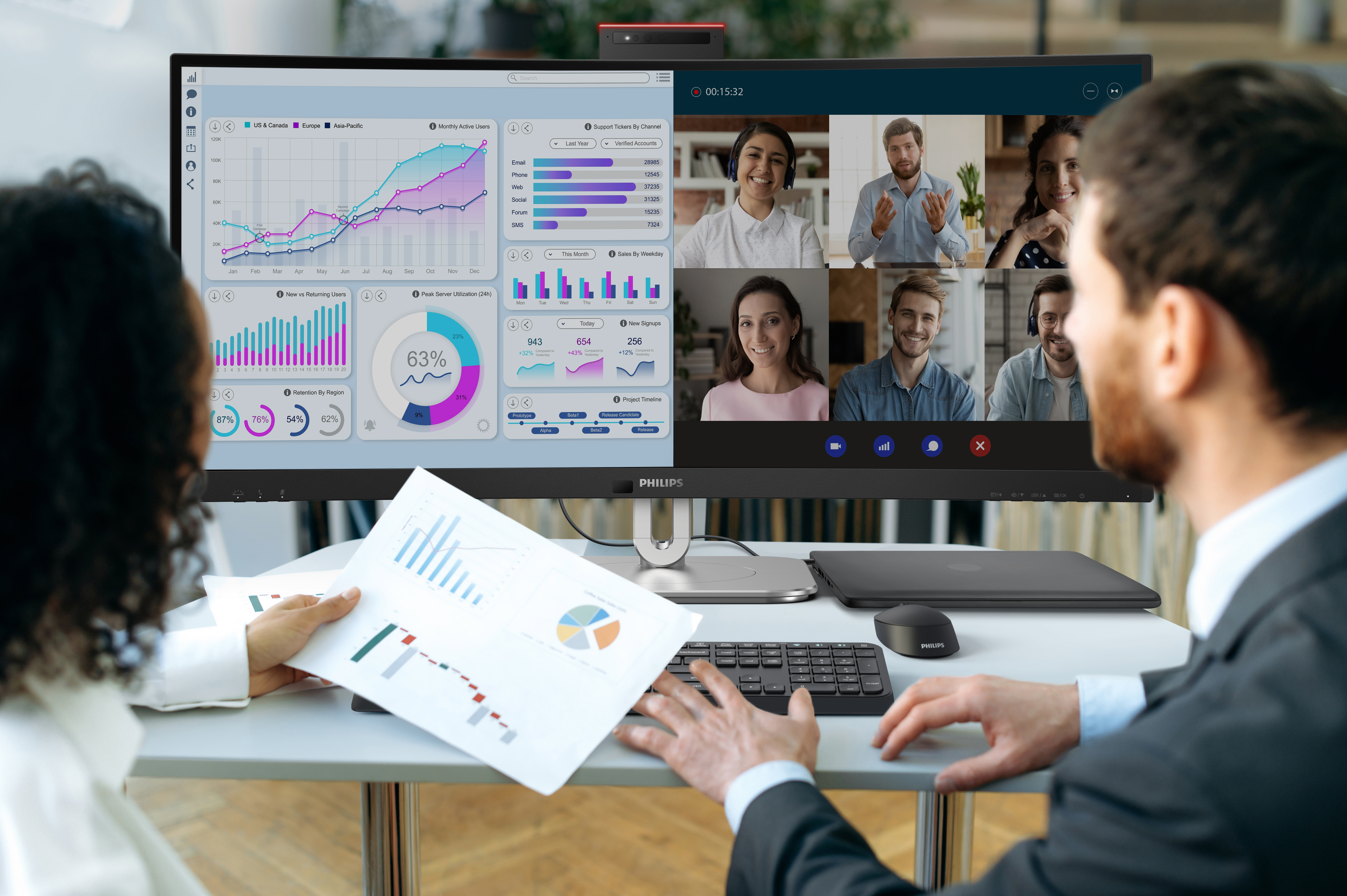Zwei Personen blicken auf einen Monitor mit einer Videokonferenz und Diagrammen.