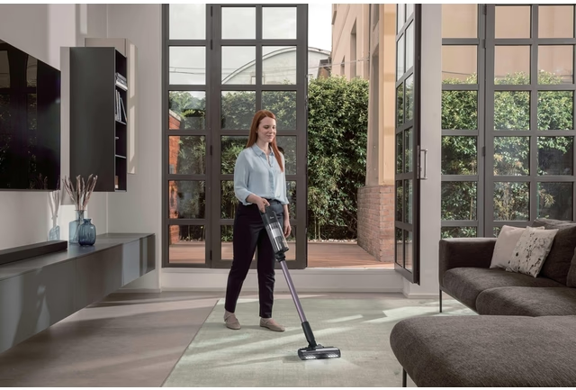 Donna aspira tappeto in soggiorno moderno con grandi finestre.