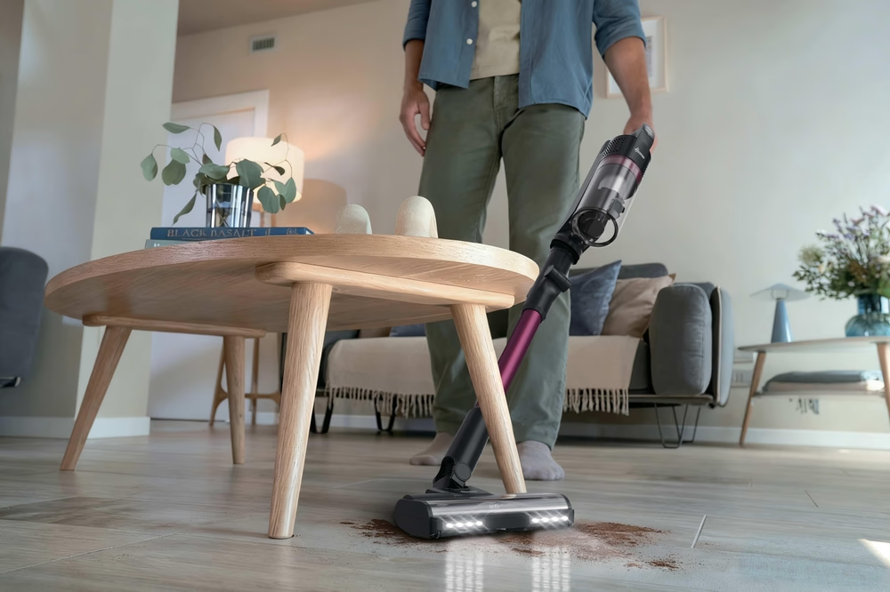 Ein Mann saugt mit einem Handstaubsauger auf dem Boden auf, der neben einem Holztisch liegt.
