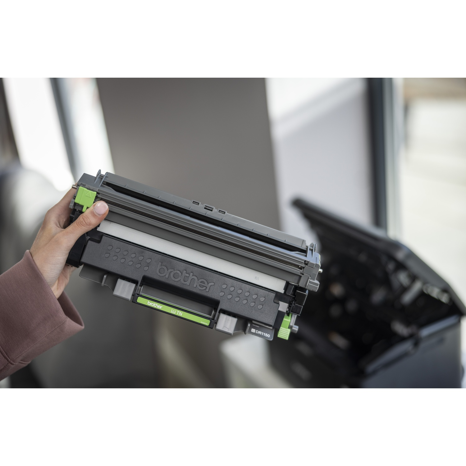 Una mano tiene una cartuccia del toner Brother con accenti verdi davanti a una stampante.