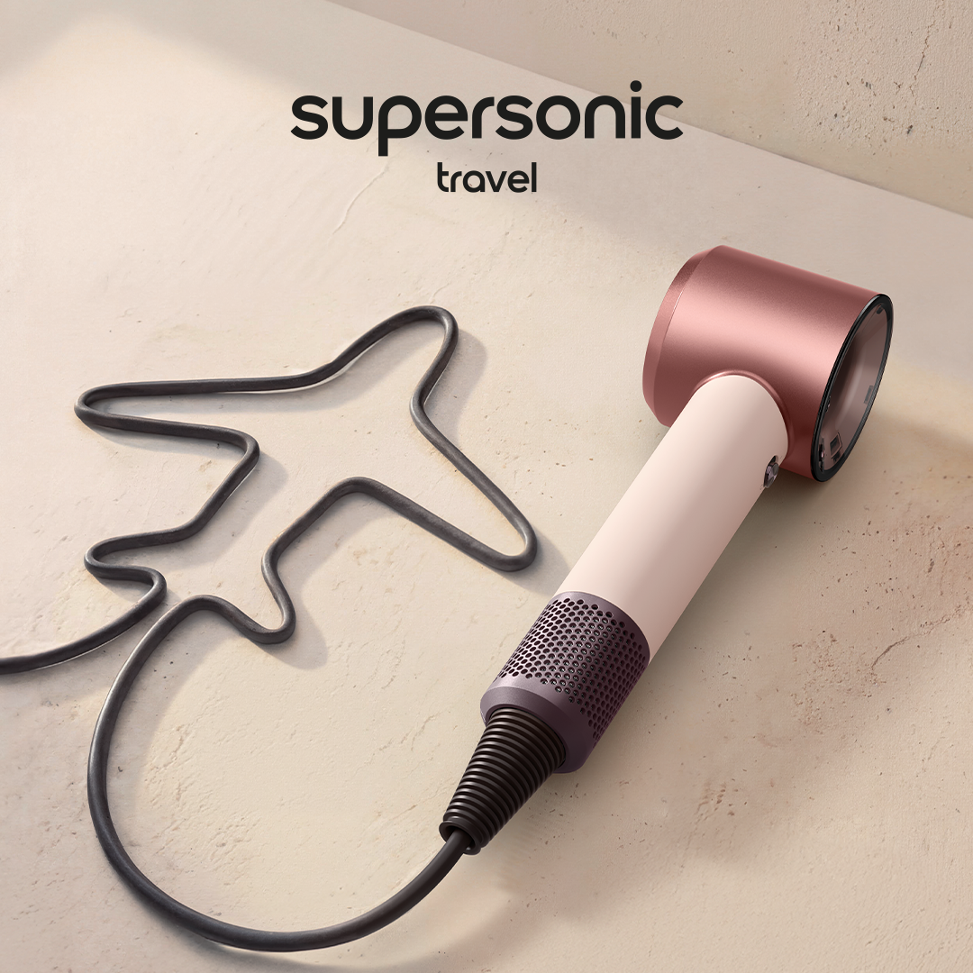 Un secador de pelo de viaje Supersonic travel color Ceramic Pink con el cable en forma de avión sobre superficie beige. Logo de Dyson en negro.