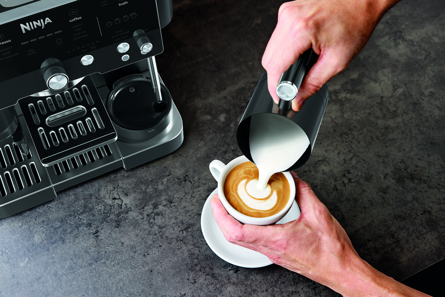 Una persona vierte leche espumada en una taza de café, creando arte latte. Cerca hay una máquina de café Ninja.