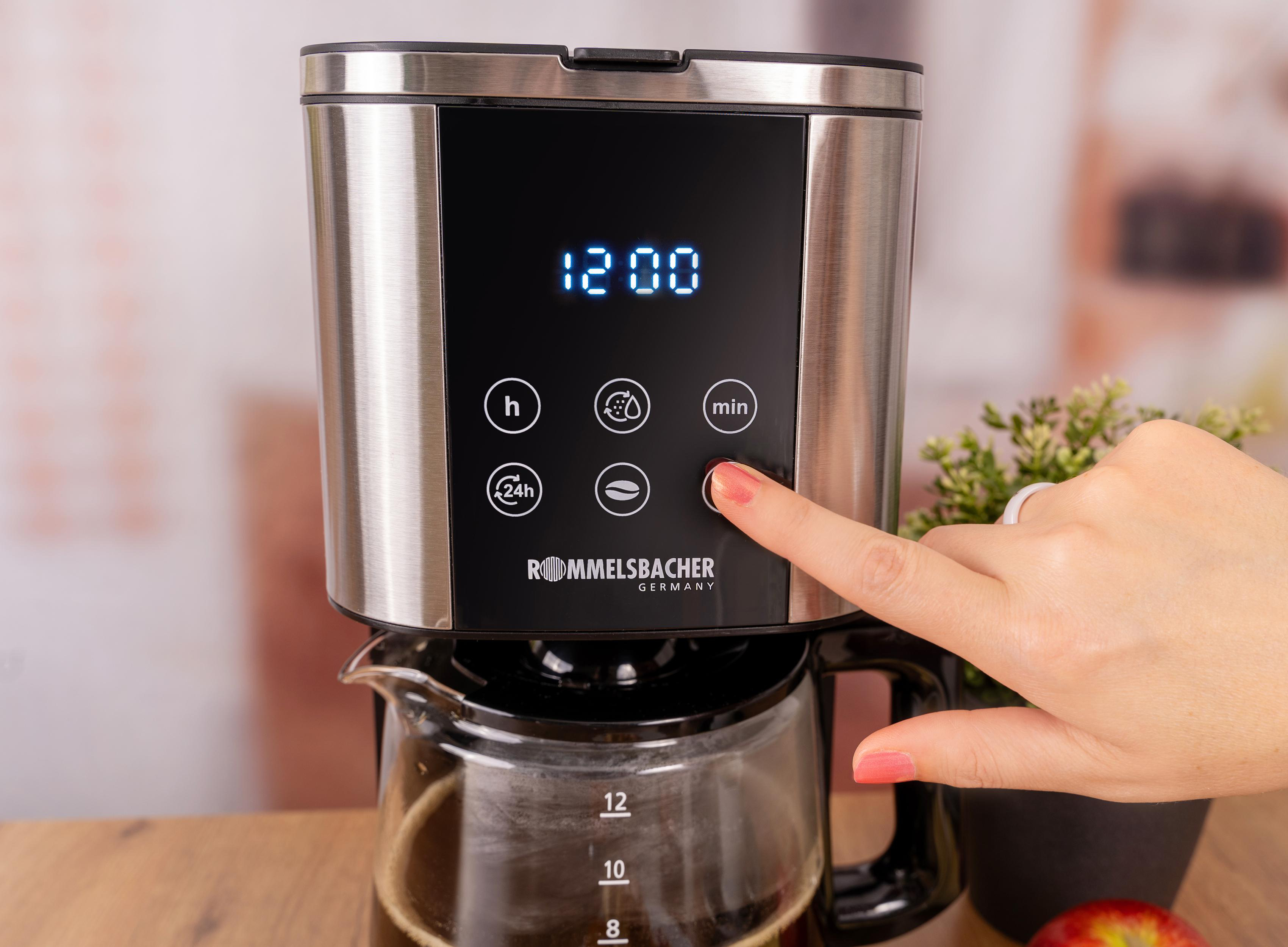 Eine Hand drückt einen Knopf an einer silbernen Kaffeemaschine mit digitaler Anzeige.