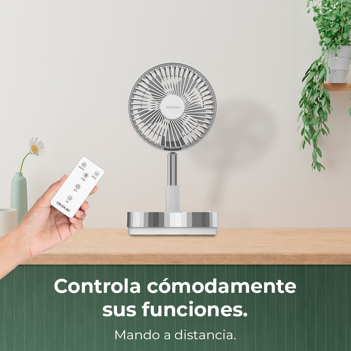 Una mano sostiene un mando a distancia junto a un ventilador blanco. Plantas al fondo.