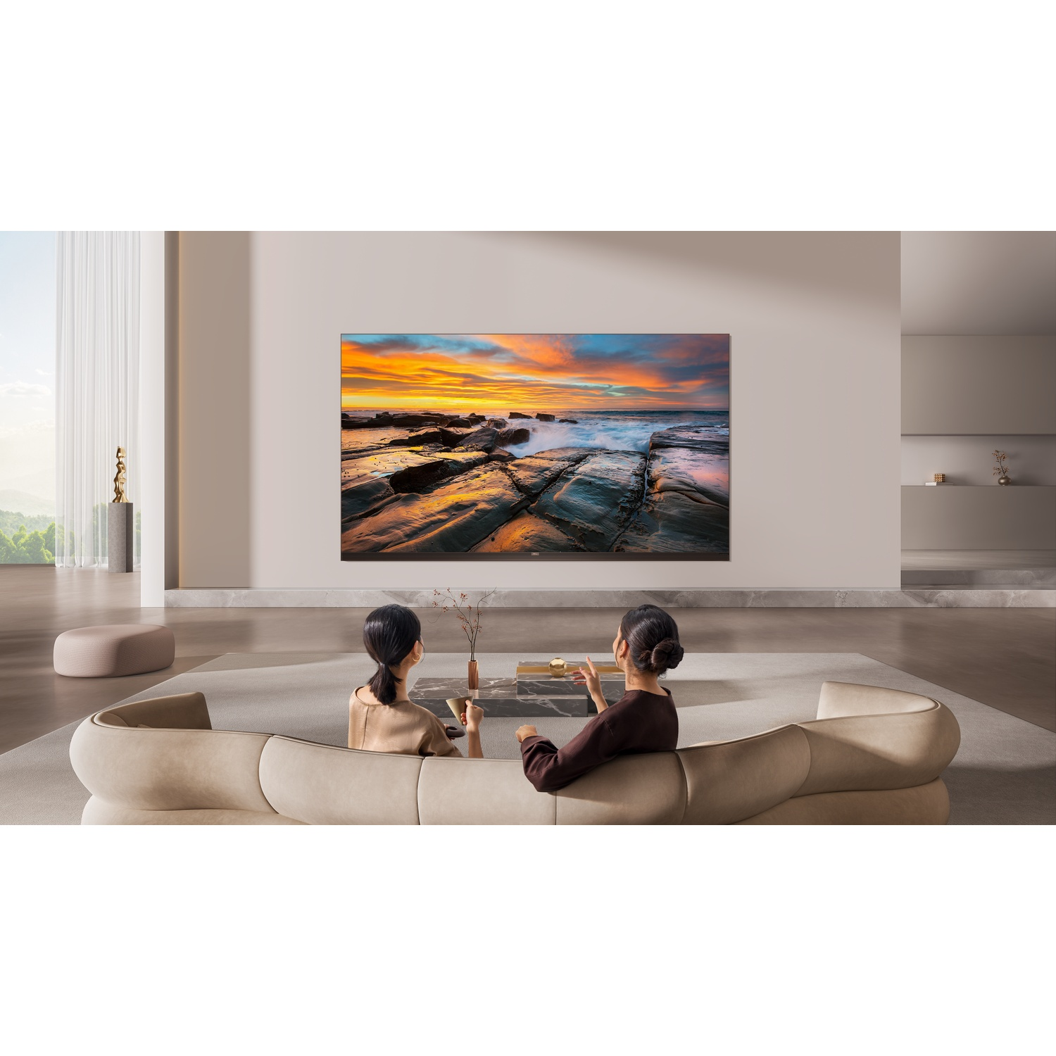 Two women on a sofa watch a large TV displaying a sunset over rocky shores.